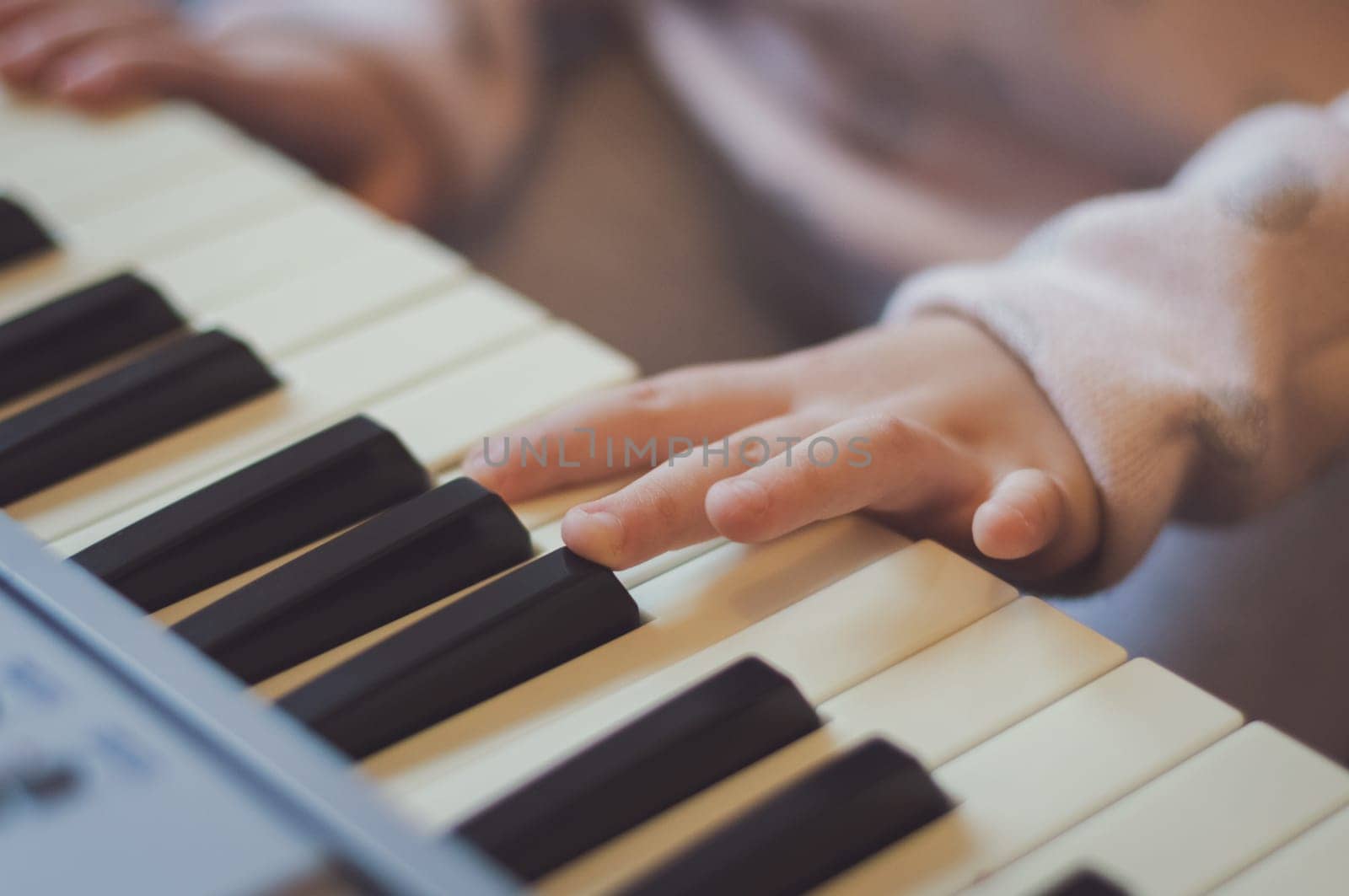 The hands of a little caucasian girl presses the keys on an electric piano while sitting on a sofa in the room, close-up side view. Music education concept.