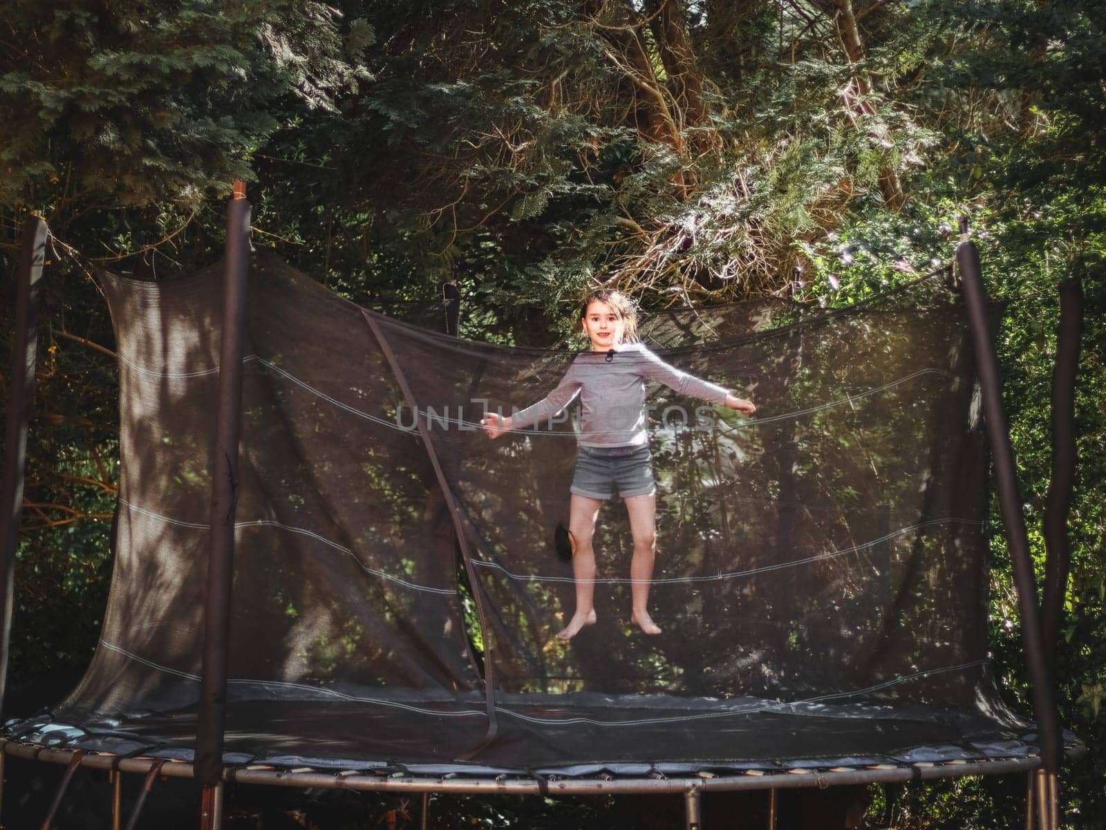 A beautiful caucasian girl in shorts and a sleeved T-shirt jumps on a trampoline, frozen in a straight position with outstretched arms in the garden of a house among trees, close-up side view. Concept at home, home sports, happy childhood, children entertainment.