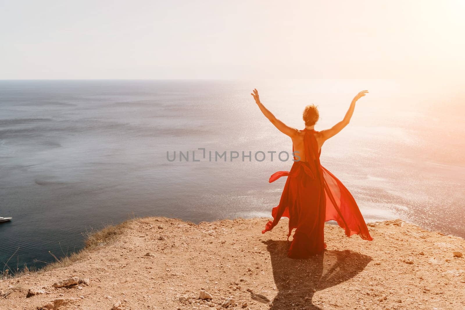 Woman in red dress on sea. Side view a Young beautiful sensual woman in a red long dress posing on a rock high above the sea on sunset. Girl on the nature on blue sky background. Fashion photo. by panophotograph