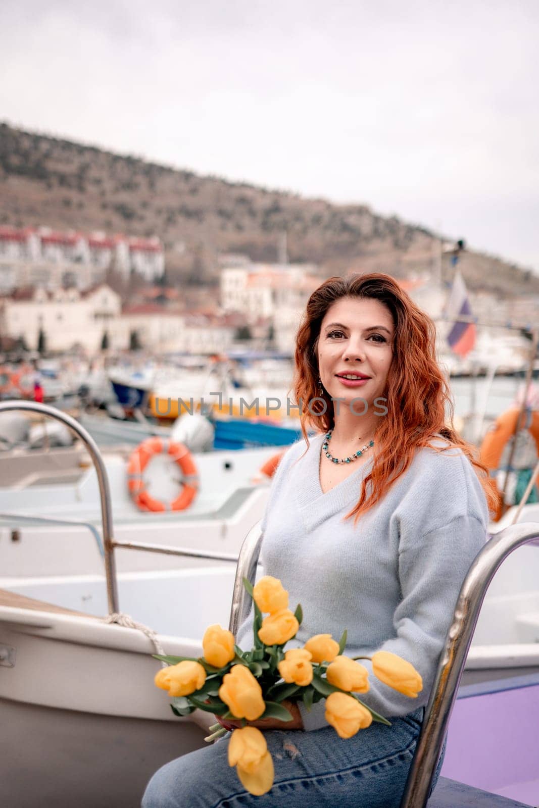 Woman holds yellow tulips in harbor with boats docked in the background., overcast day, yellow sweater, mountains.