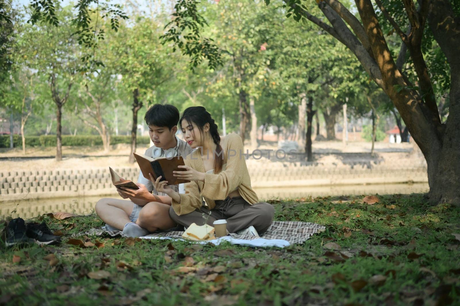 Relaxed young couple spending time together outdoor reading book on picnic blanket in the park.