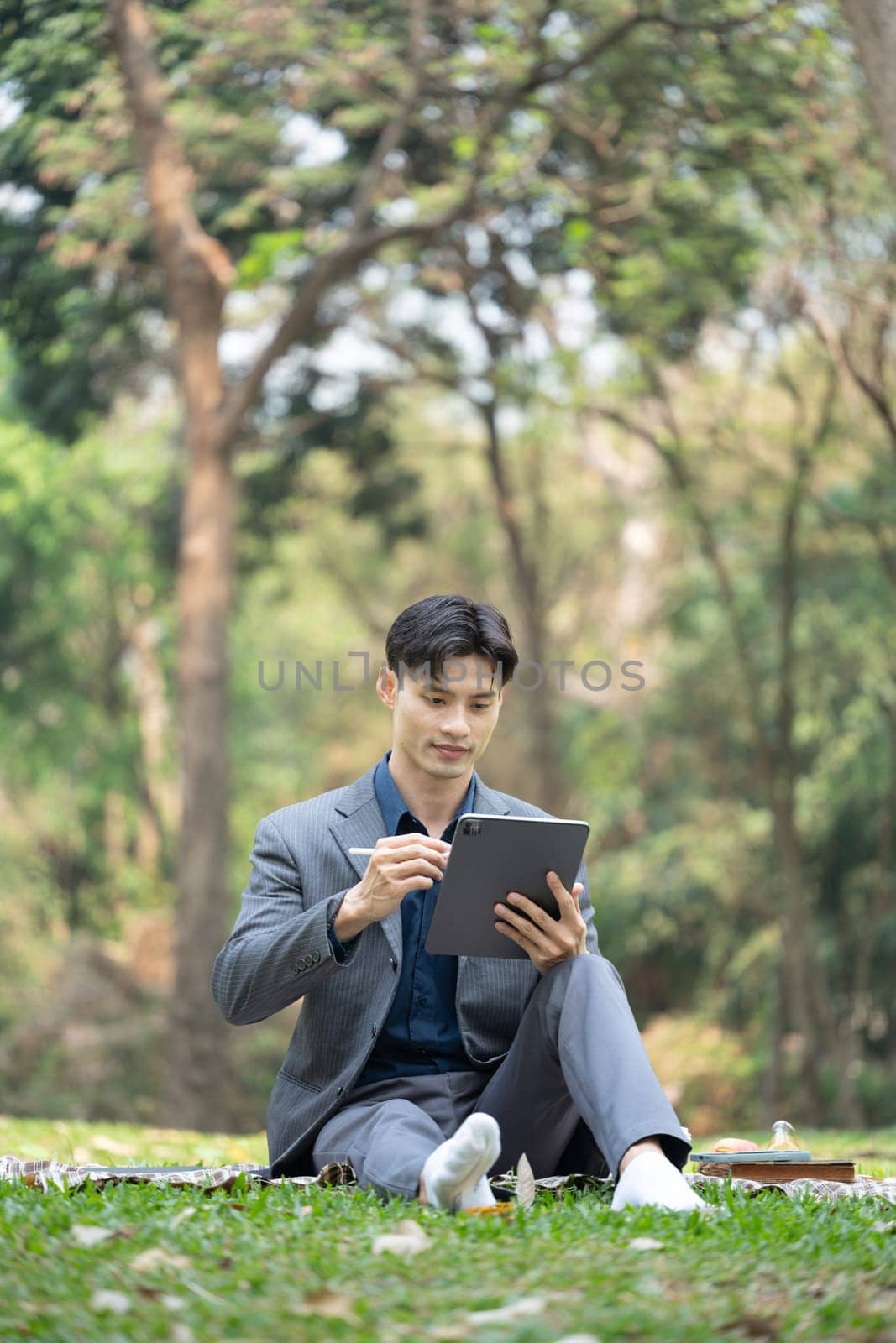 Portrait of businessman sitting on green grass in the city park and using digital tablet.