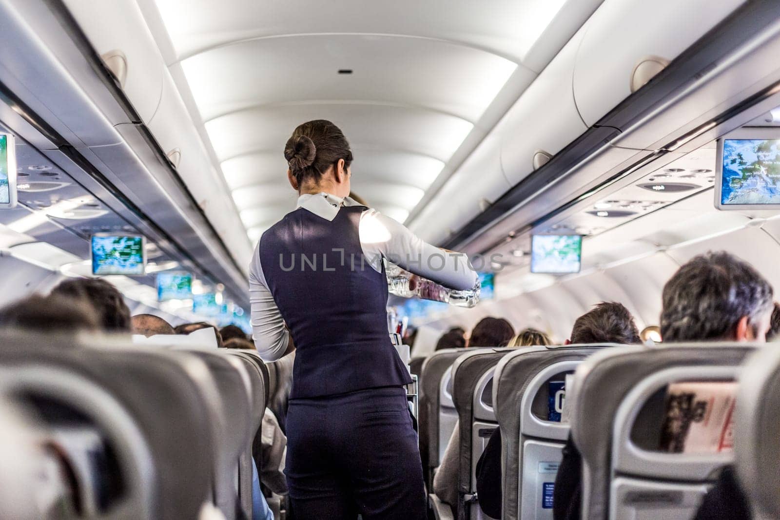 Interior of airplane with passengers on seats and stewardess in uniform walking the aisle, serving people. Commercial economy flight service concept