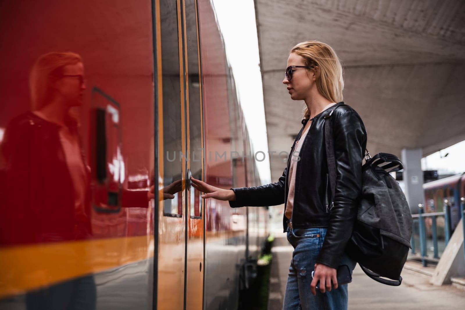 Young blond woman in jeans, shirt and leather jacket wearing bag and sunglass, presses door button of modern speed train to embark on train station platform. Travel and transportation. by kasto