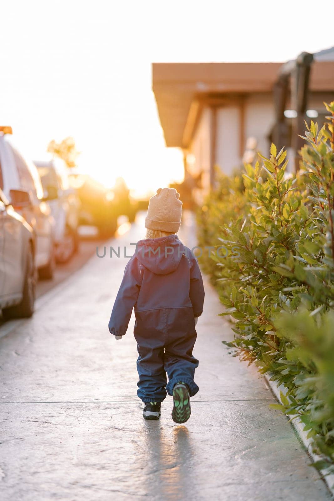 Little girl walks along a sunny sidewalk along parked cars. Back view. High quality photo