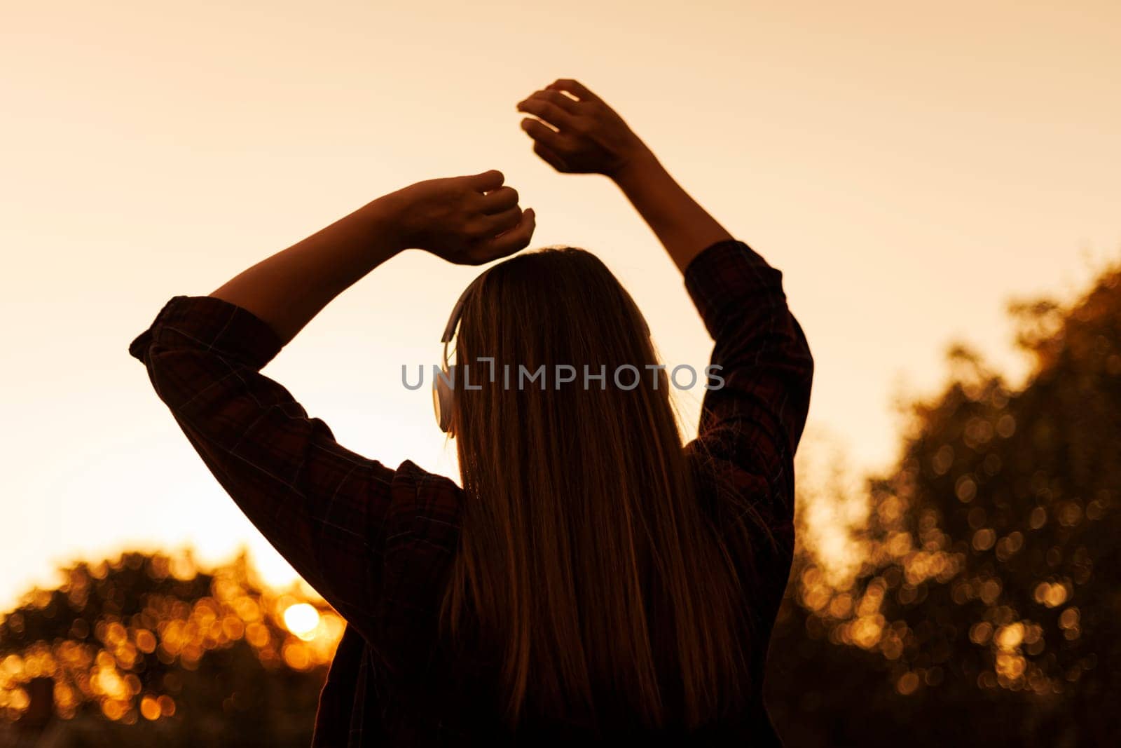 Silhouette of a young woman with headphones enjoying music at sunset, with trees in the background. Casual lifestyle photography. Relaxation and leisure concept. Design for poster, banner, wallpaper.