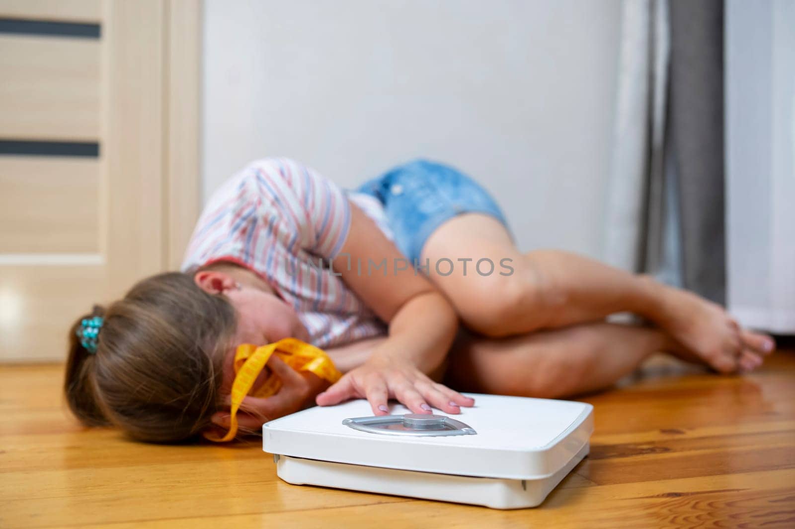 Distressed girl lying on the floor next to a weighing scale and measuring tape by andreyz