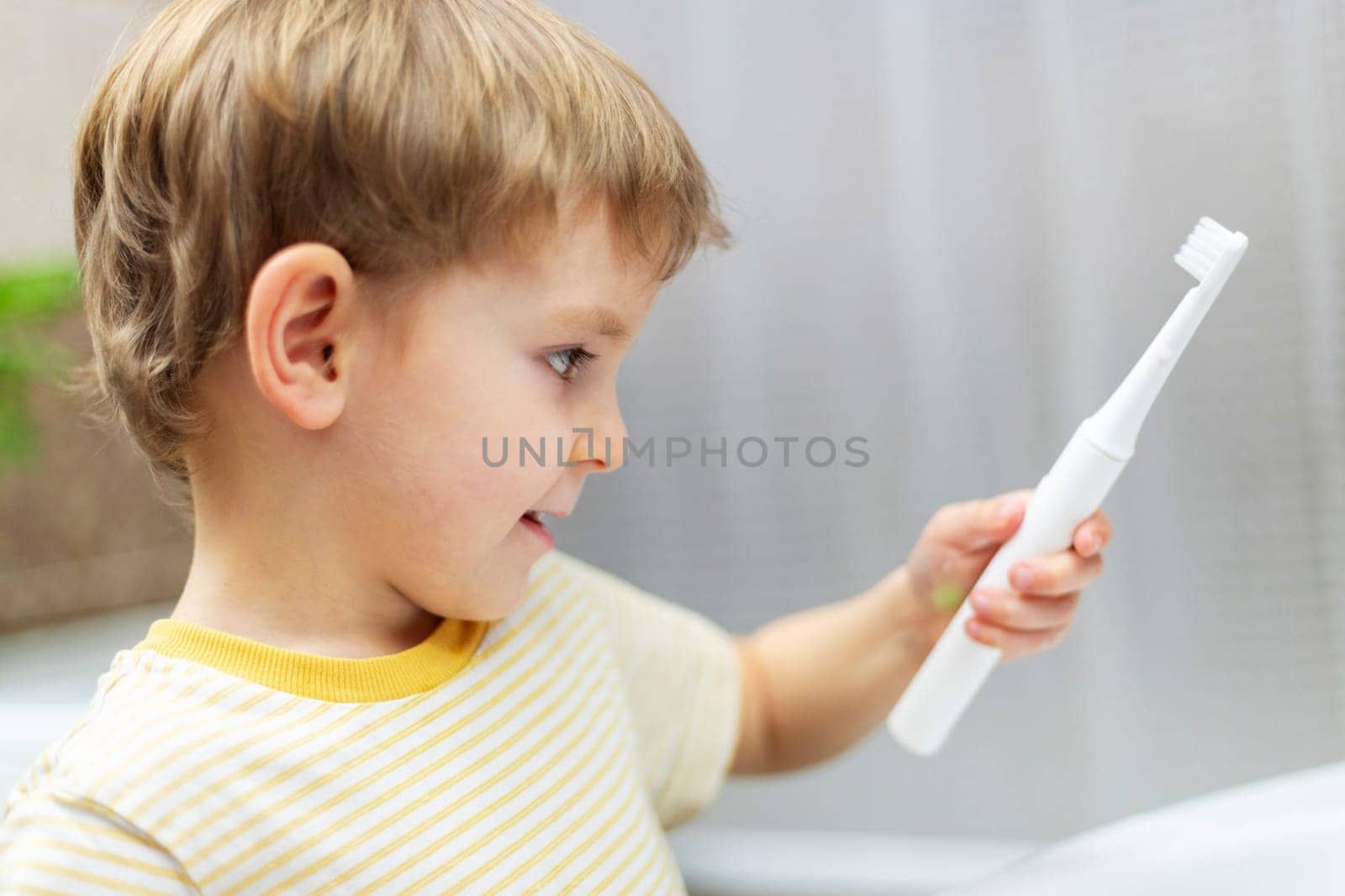 Smiling boy holding electric toothbrush, looking at it curiously in bathroom. Oral hygiene and health care concept for children. Design for educational poster, banner, and healthcare promotion.