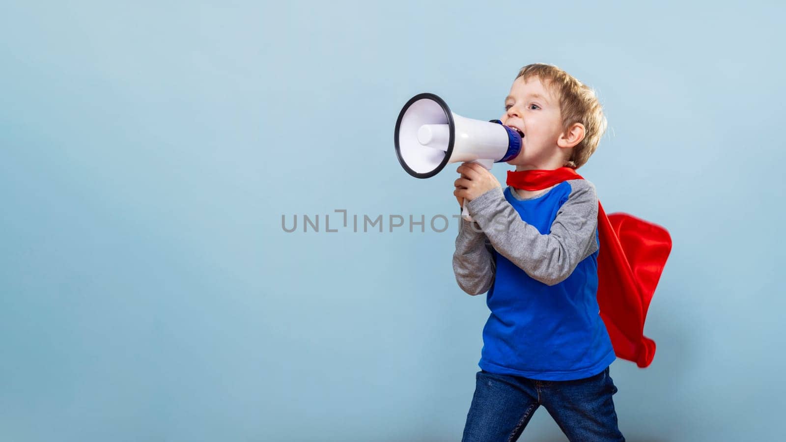 Child in superhero costume with red cape shouting into megaphone against blue background. Kid imagination and play concept. Design for banner, poster, invitation with copy space.