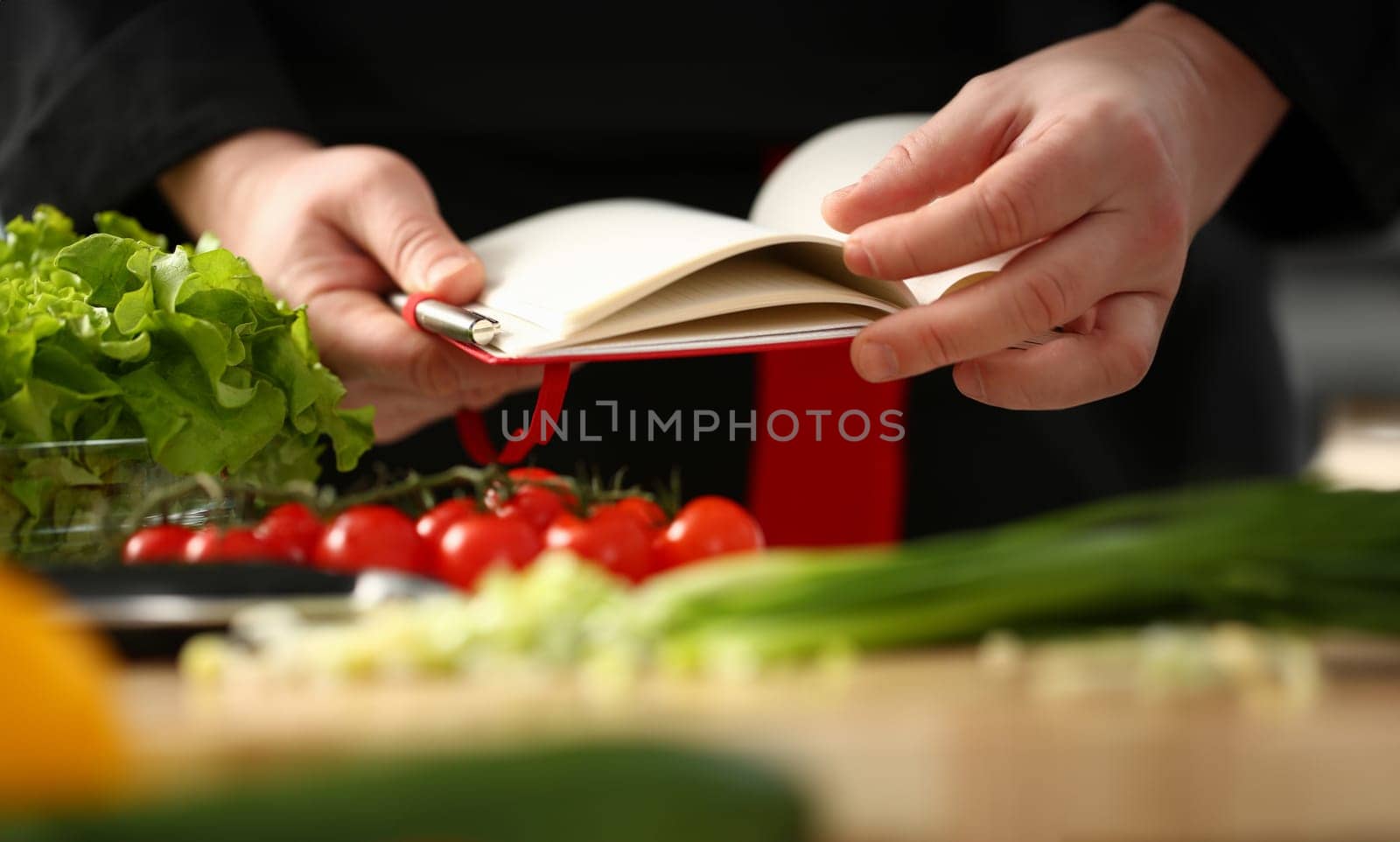 Male cook hand hold siver pen and wright open book of recipes on background of vegetables. Chef writes new combination of taste in home magazine shopping organizer concept list