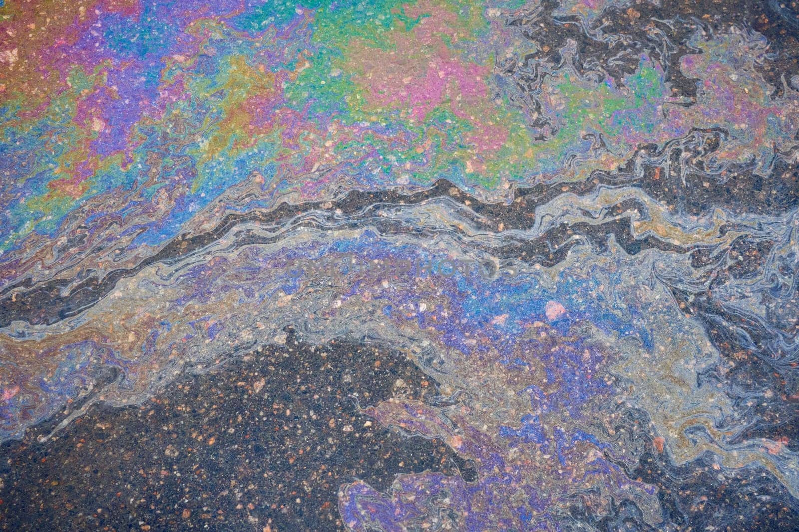 Oil residues on wet asphalt interact with sunlight, displaying a rainbow-like spectrum of colors