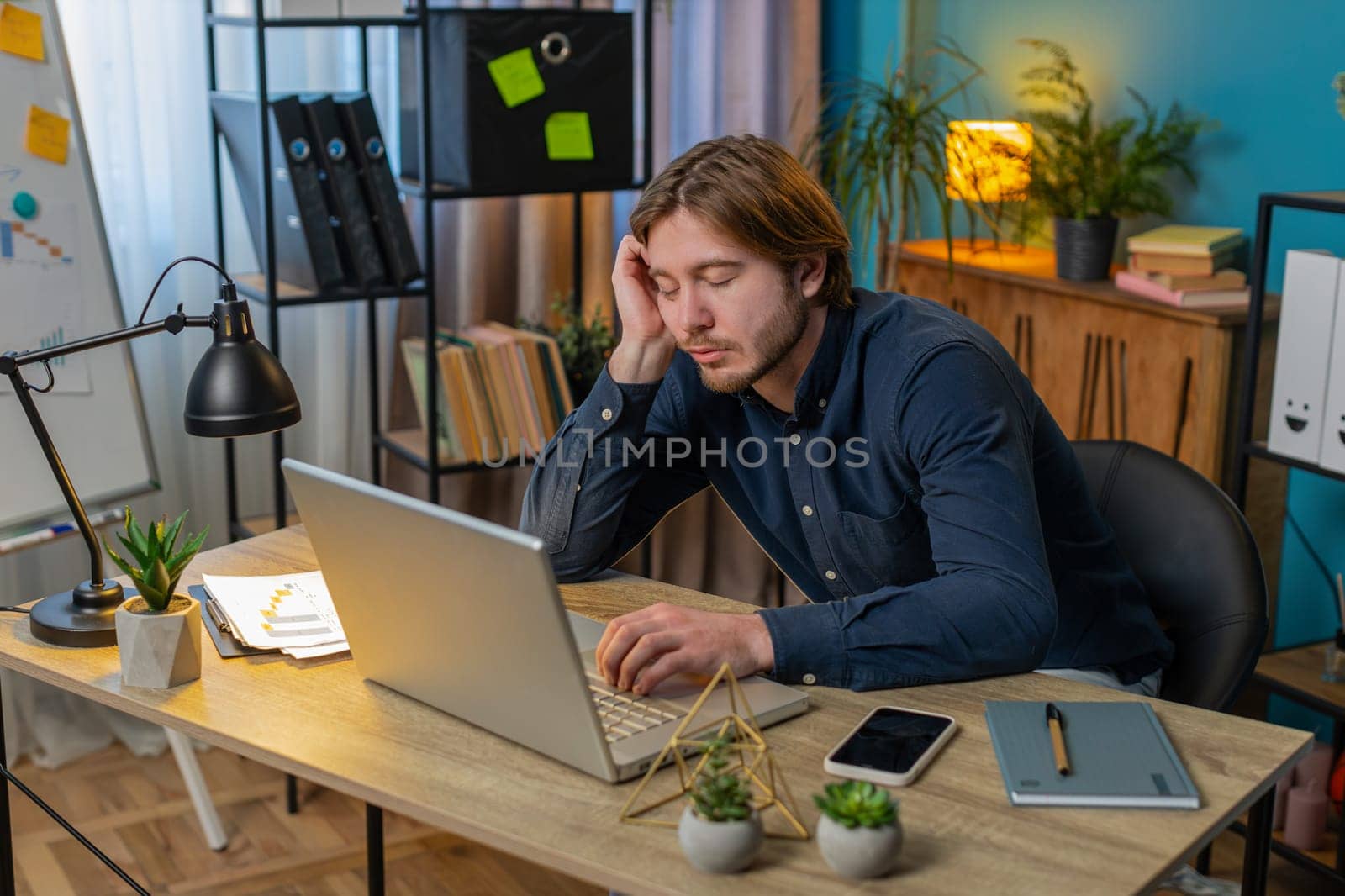 Bored sleepy businessman worker working on laptop computer leaning on hand sleeps at home office desk. Exhausted tired Caucasian young man freelancer workaholic guy. Employment, occupation, workless.