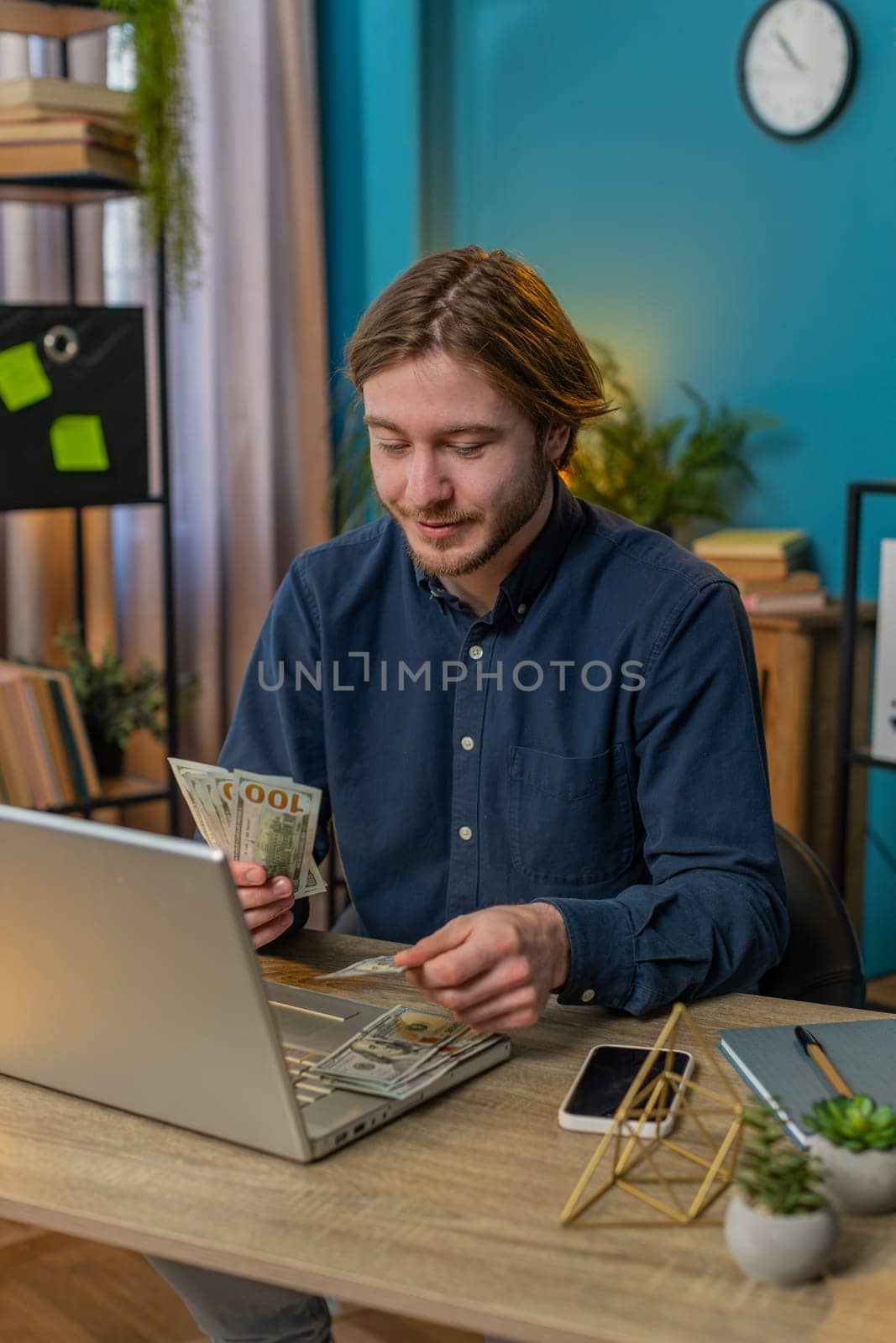 Young businessman guy holding fan of cash money dollar banknotes showing thumbs up and pointing at laptop, celebrate, success career, online income salary wealth at home office. Remote distant working
