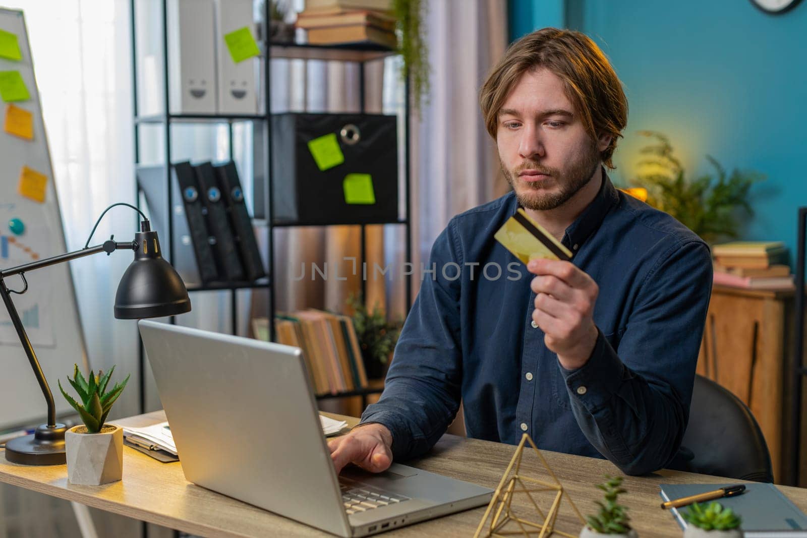 Young Caucasian businessman using bank credit card and laptop for online shopping payments at home office. Business transaction. Strict guy freelancer purchases with netbook E-banking app service.