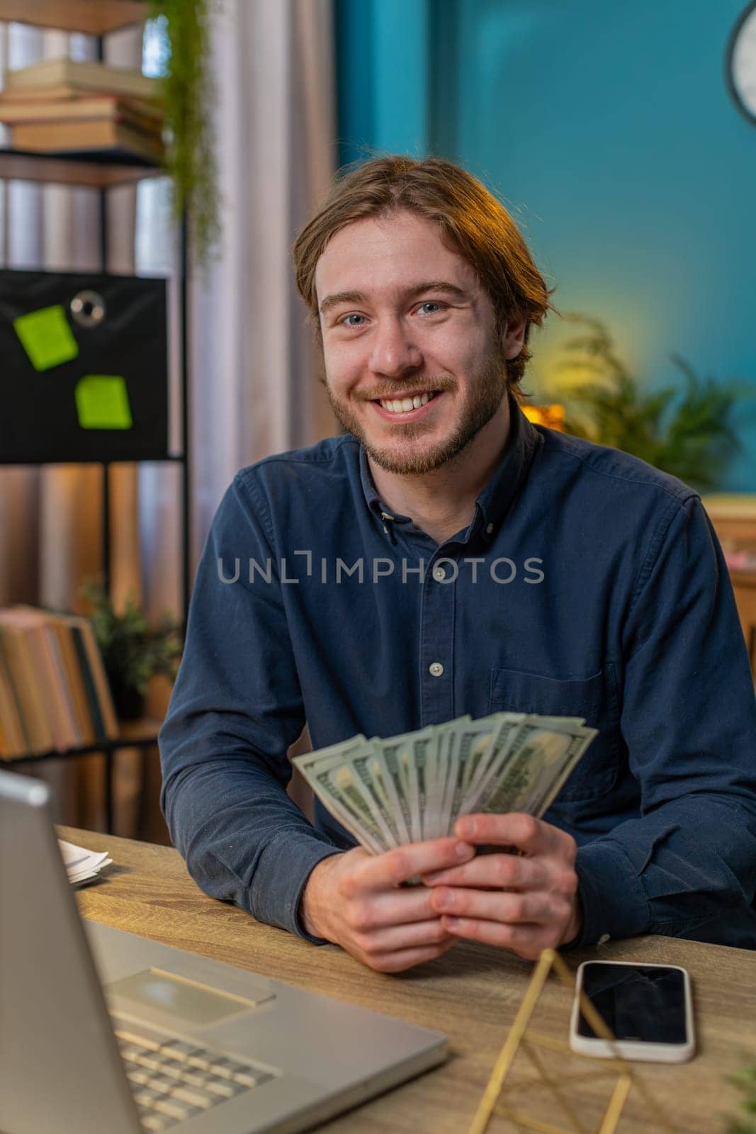 Young businessman guy holding fan of cash money dollar banknotes showing thumbs up and pointing at laptop, celebrate, success career, online income, wealth at home office table. Remote distant working
