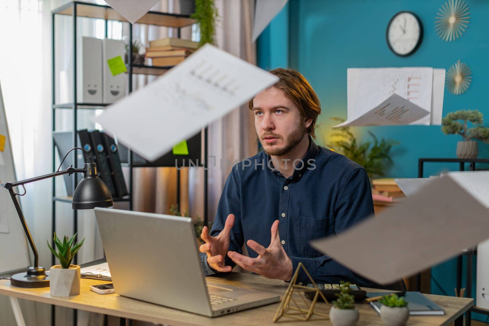 Frustrated upset young businessman works at home office desk looking at bills paperwork reports documents gets angry and throws documents in air, financial problem. Stresses freelancer guy at home.