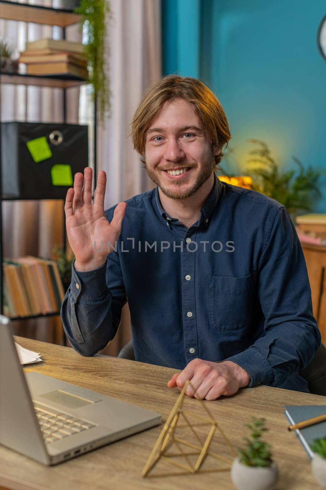 Hello. Happy businessman employee waves hand hi gesture greeting welcomes someone, online webinar, remote distance conversation. Freelance man sitting at desk using laptop in home office. Vertical