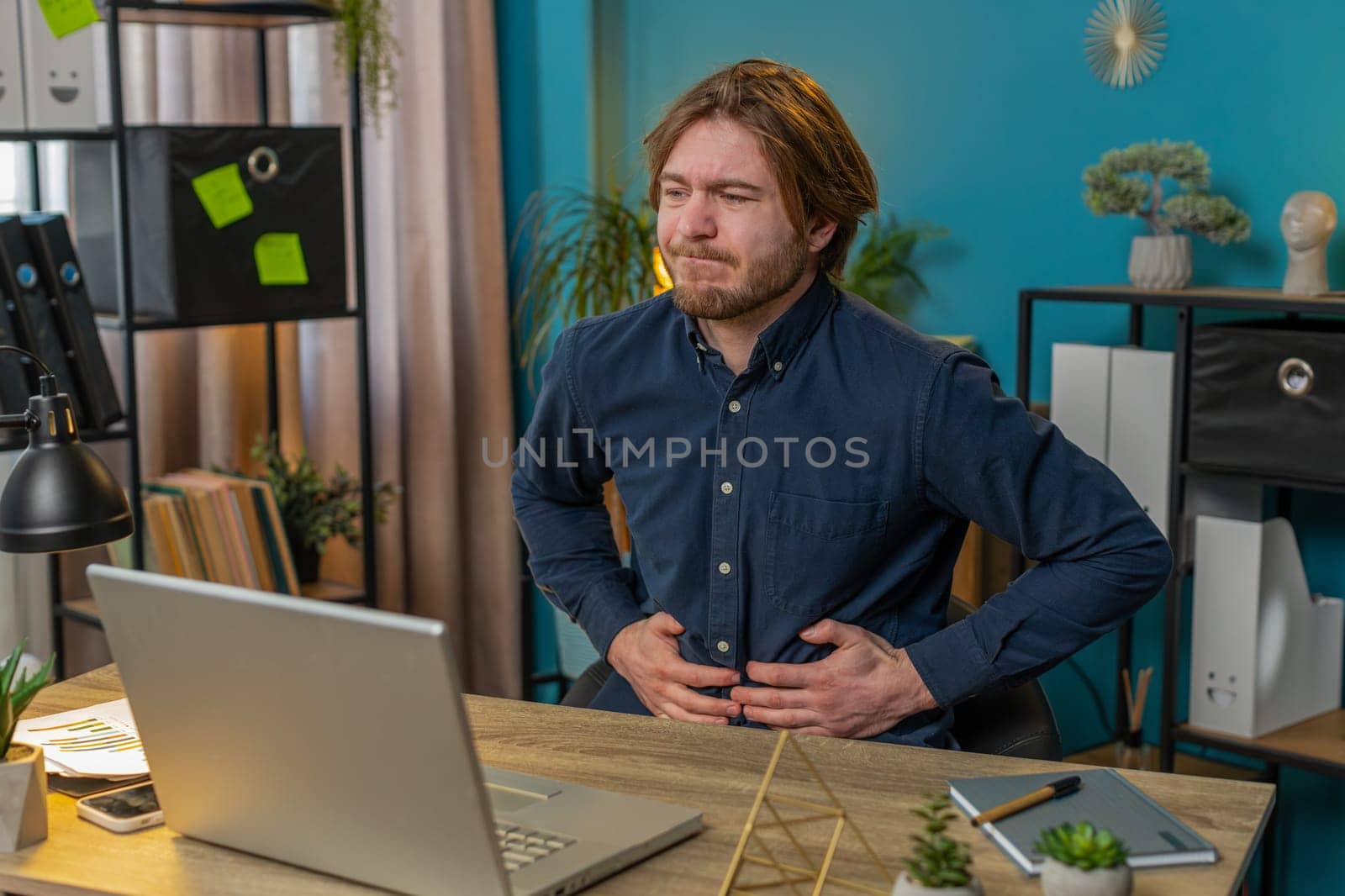 Disappointed Caucasian freelancer feeling overeating during work process on laptop in home office desk. Upset businessman freelancer guy man touching stomach and experiencing discomfort and sharp pain