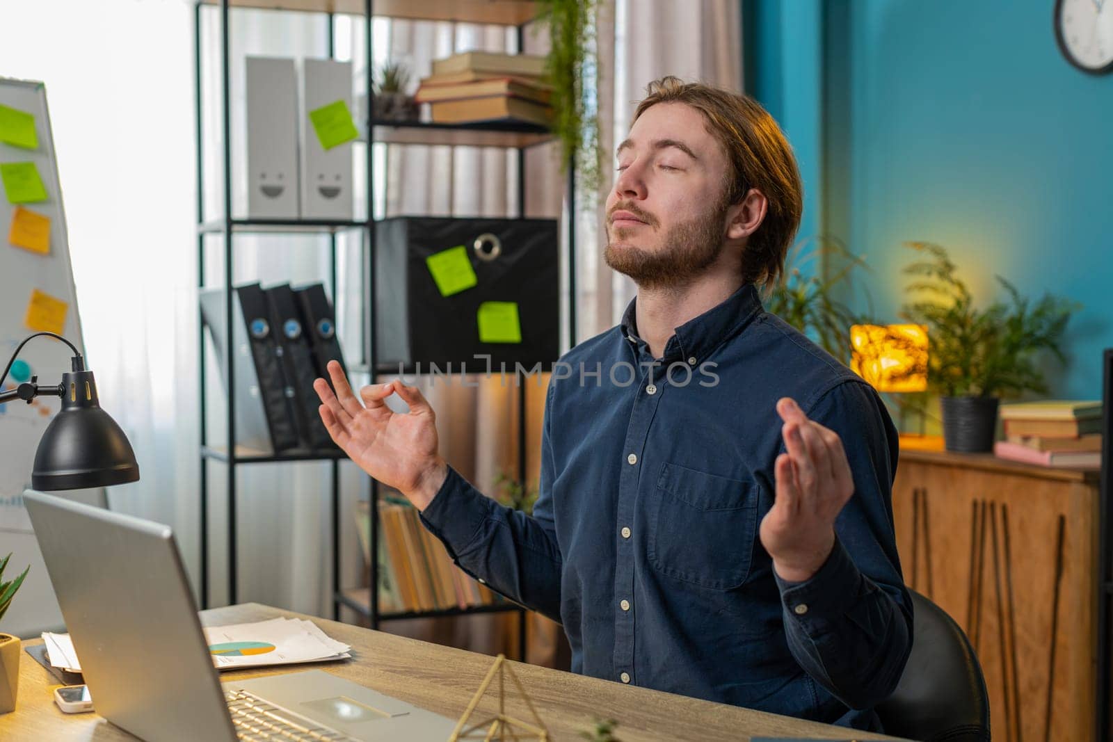 Peaceful relaxed young male worker businessman sitting at workplace with folded in mudra gesture hands, reducing stress during workday, meditating or doing yoga breathing exercises in home office