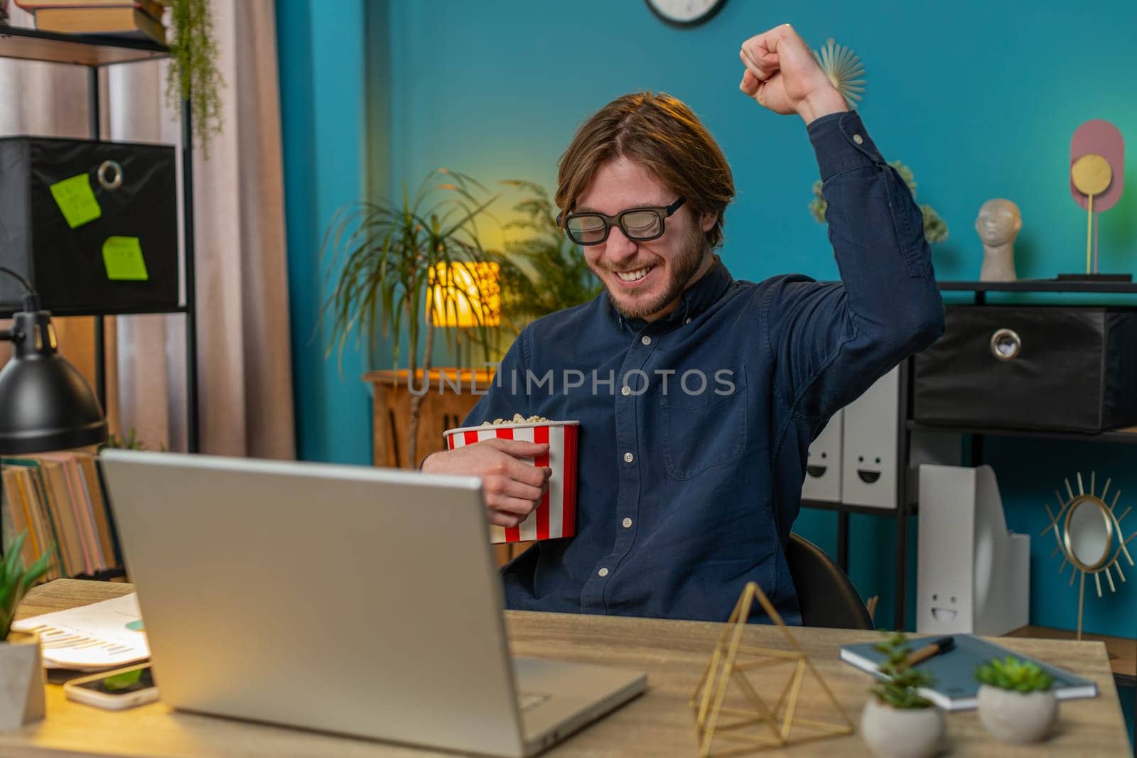 Excited happy businessman freelancer taking break from work wearing 3D glasses eating popcorn and watching movie on laptop. Laughing young man sitting at home office desk having snacks during break.