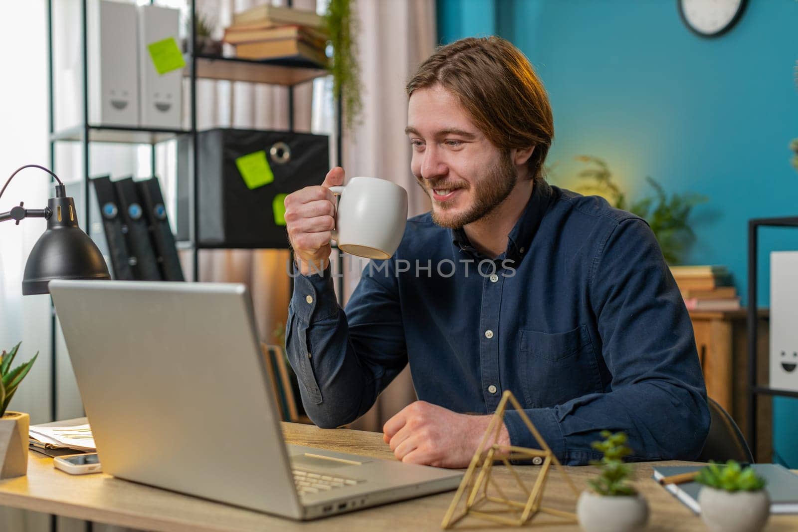 Young Caucasian businessman working on laptop computer drinking cup of warm hot coffee sits at desk workplace home office room in the morning. Male freelancer guy enjoying relaxing, taking a break.