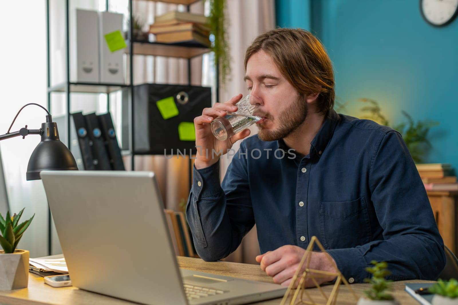 Focused Caucasian businessman sitting at workplace desk drinking water while working with laptop in home office. Young male freelancer employee man looks at computer screen browse surf internet.