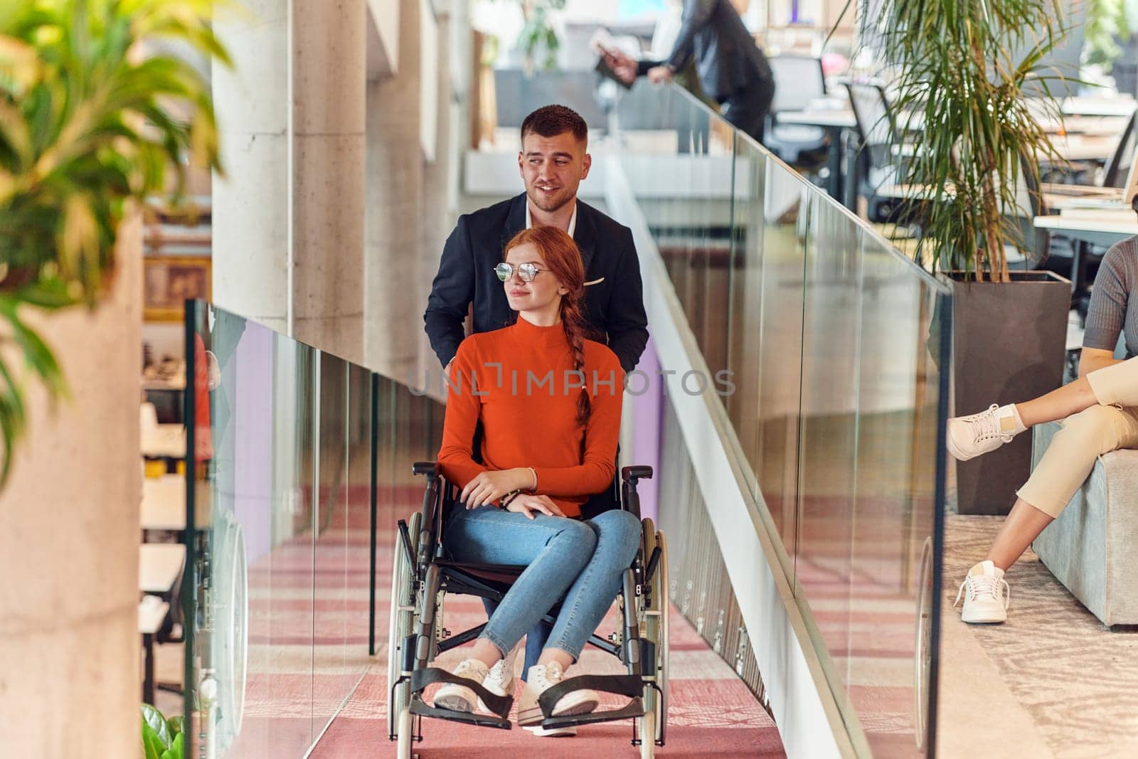 A company director assists his business colleague in a wheelchair, helping her navigate to their startup office, where they work alongside their diverse team of colleagues, emphasizing inclusivity and support in the workplace. by dotshock