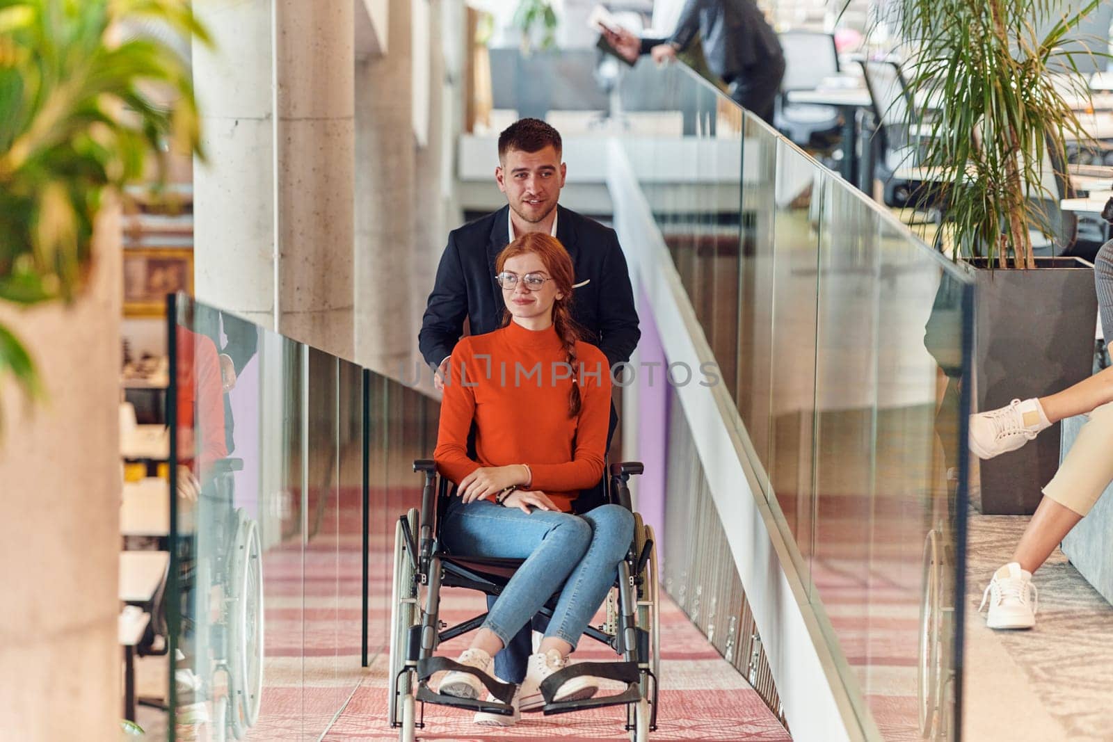 A company director assists his business colleague in a wheelchair, helping her navigate to their startup office, where they work alongside their diverse team of colleagues, emphasizing inclusivity and support in the workplace
