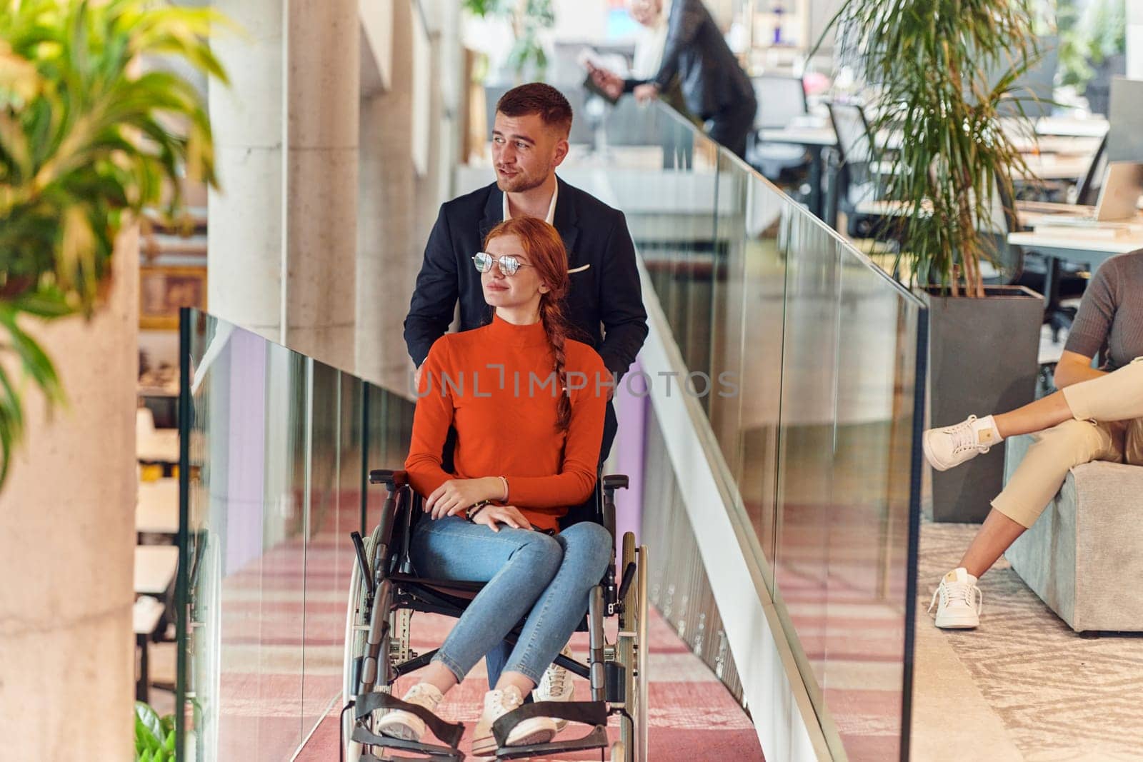 A company director assists his business colleague in a wheelchair, helping her navigate to their startup office, where they work alongside their diverse team of colleagues, emphasizing inclusivity and support in the workplace
