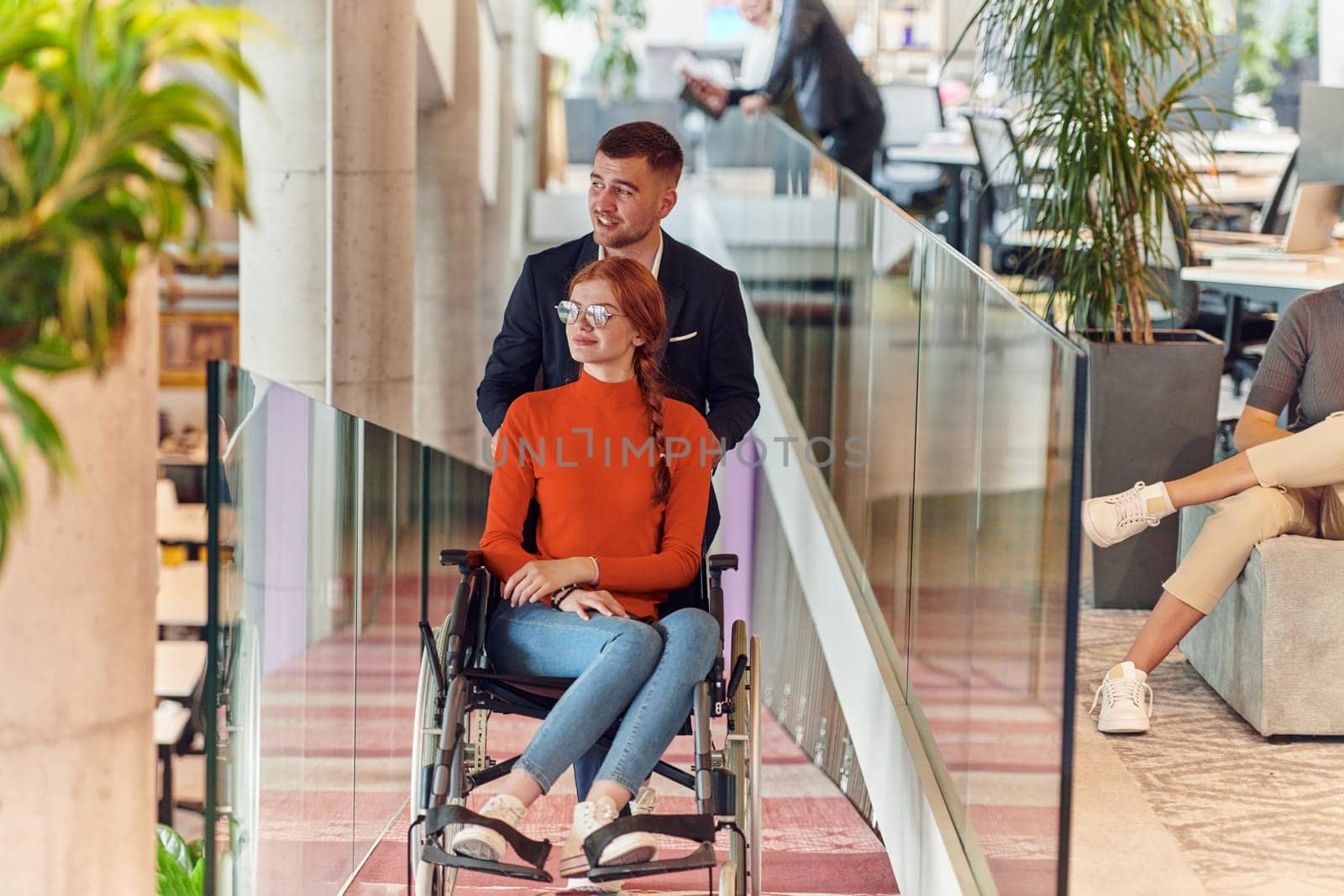 A company director assists his business colleague in a wheelchair, helping her navigate to their startup office, where they work alongside their diverse team of colleagues, emphasizing inclusivity and support in the workplace