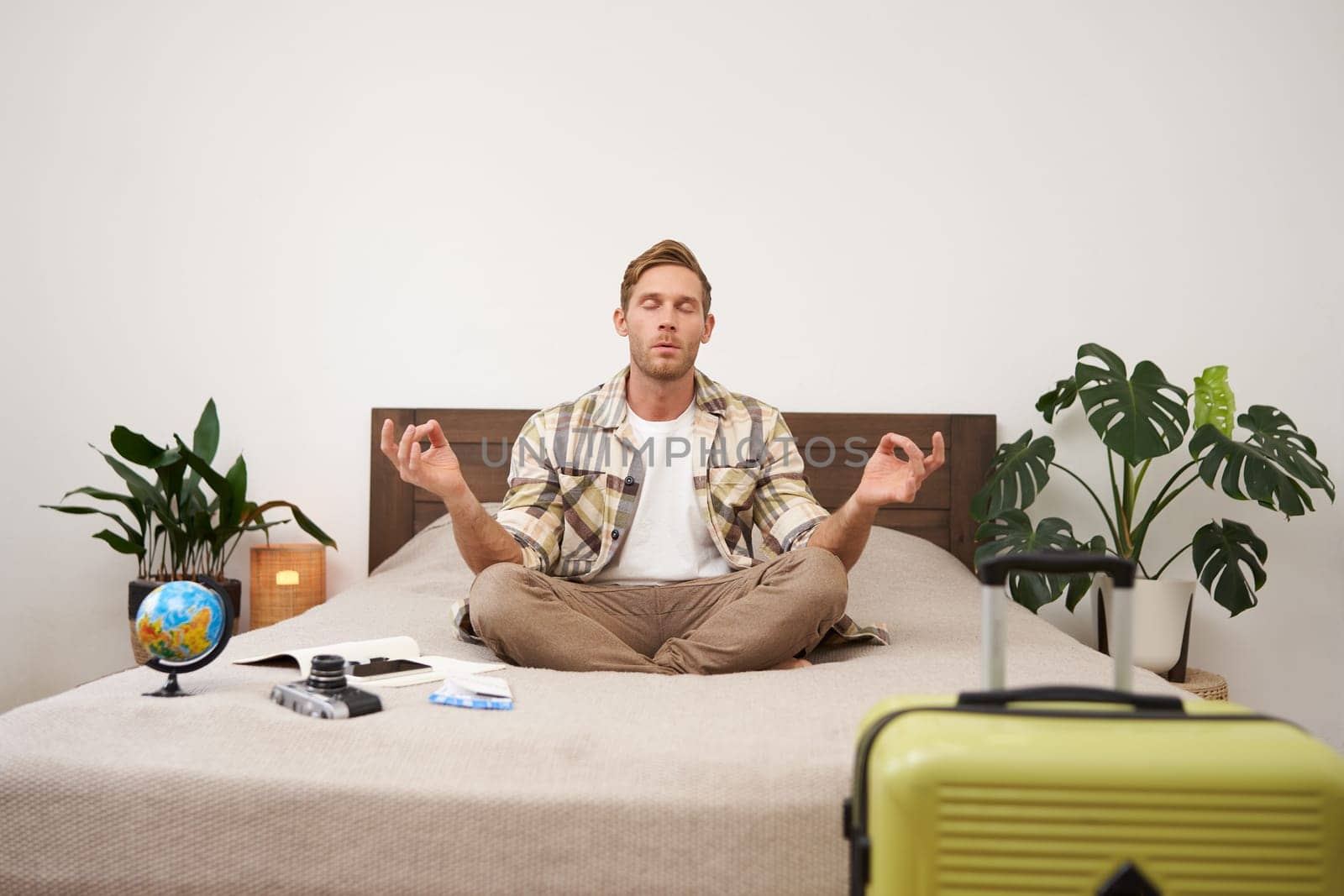 Portrait of young mindful traveller, man sitting on bed and meditating, packed suitcase, going to travel abroad, practice yoga, has a globe and camera lying next to him.