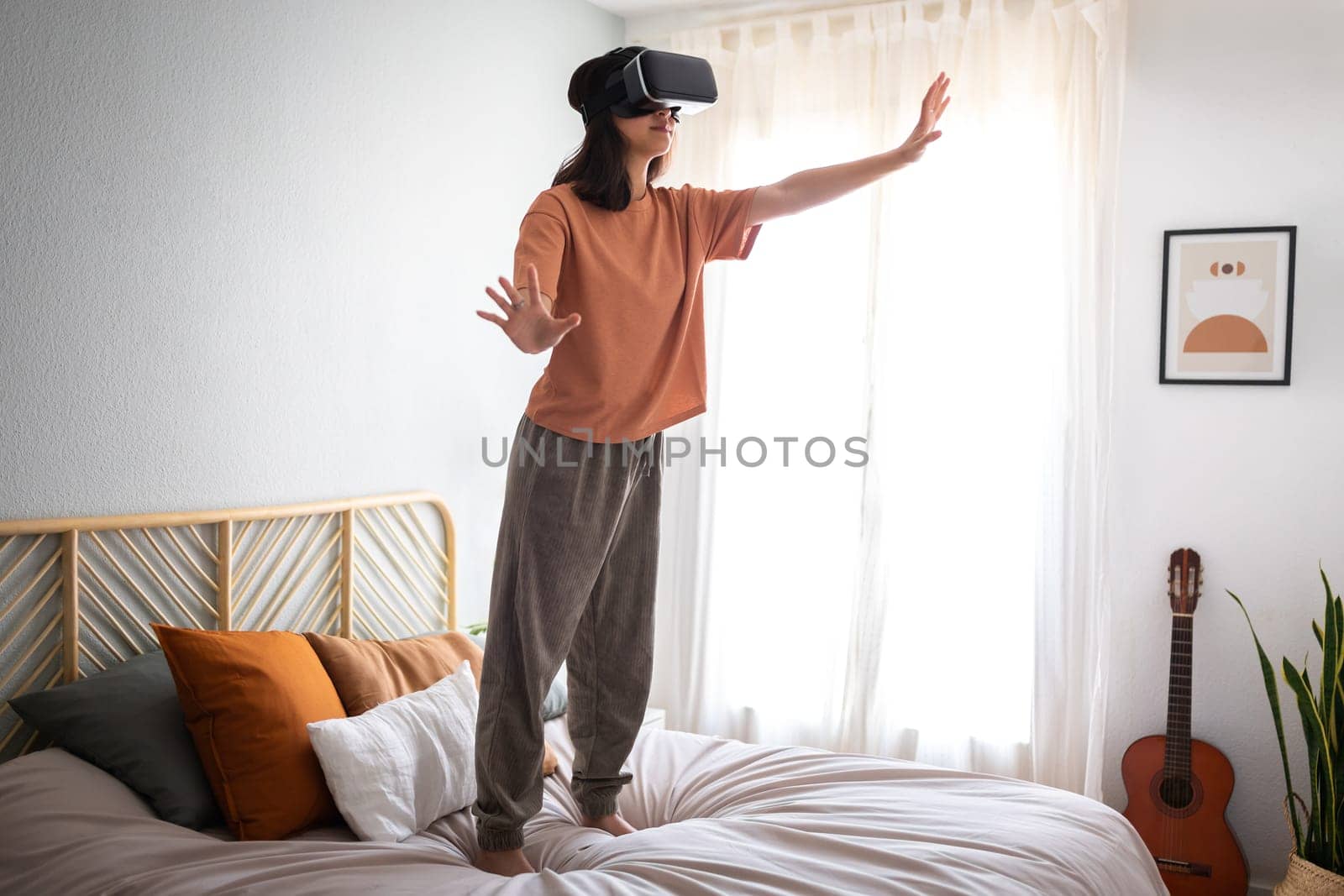 Teen girl standing on bed, moving hands touching objects in 3D world video game using VR headset. Young woman using virtual reality goggles at home bedroom. Copy space. Modern technology.