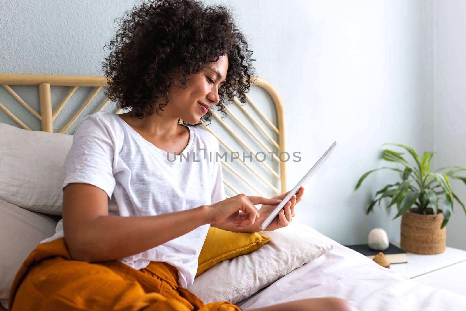 Young African American woman using tablet lying down relaxing on bed at home cozy bedroom. Technology concept.