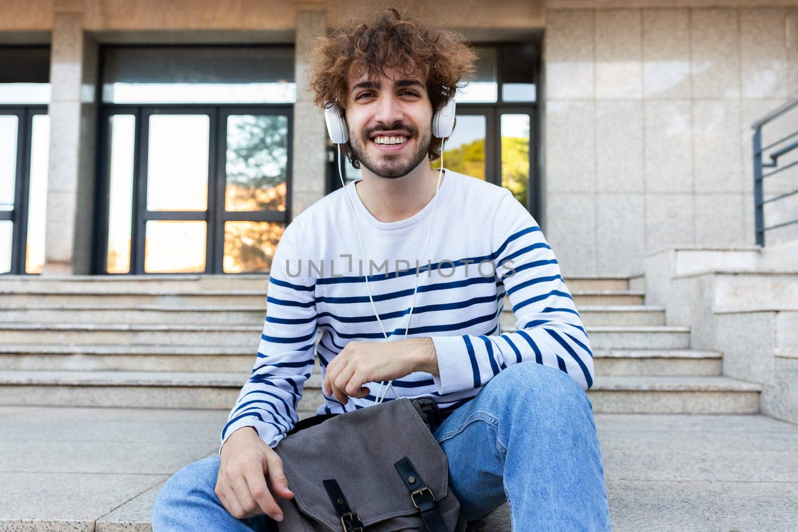 Happy young university student sitting on stairs in campus wearing headphones looking at camera. Education concept.