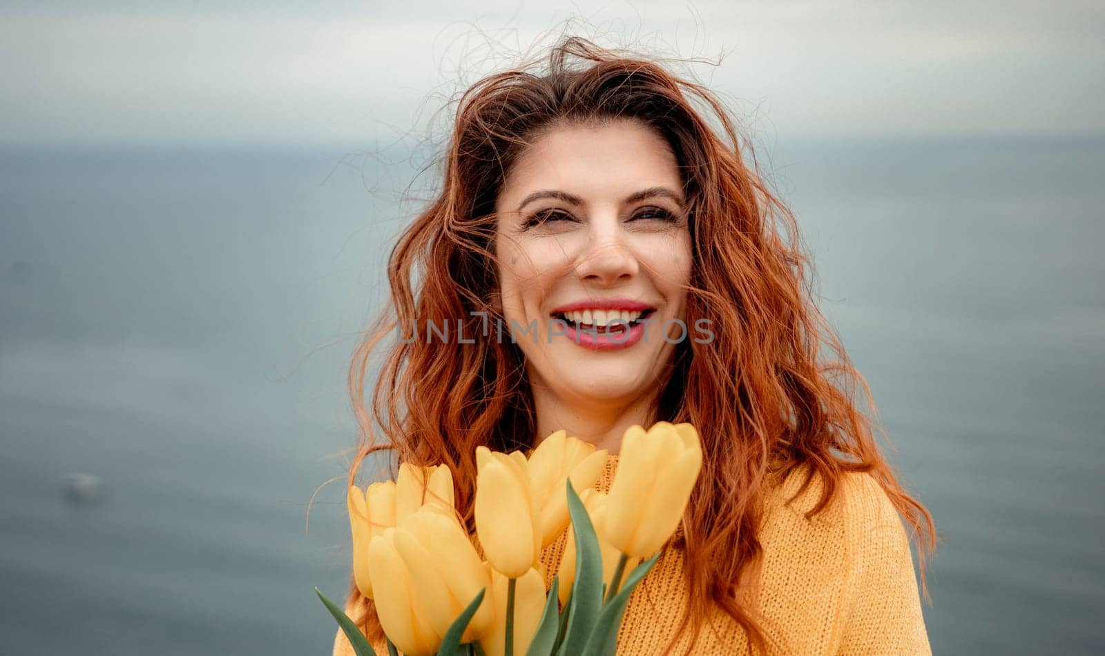 Portrait of a happy woman with hair flying in the wind against the backdrop of mountains and sea. Holding a bouquet of yellow tulips in her hands, wearing a yellow sweater.