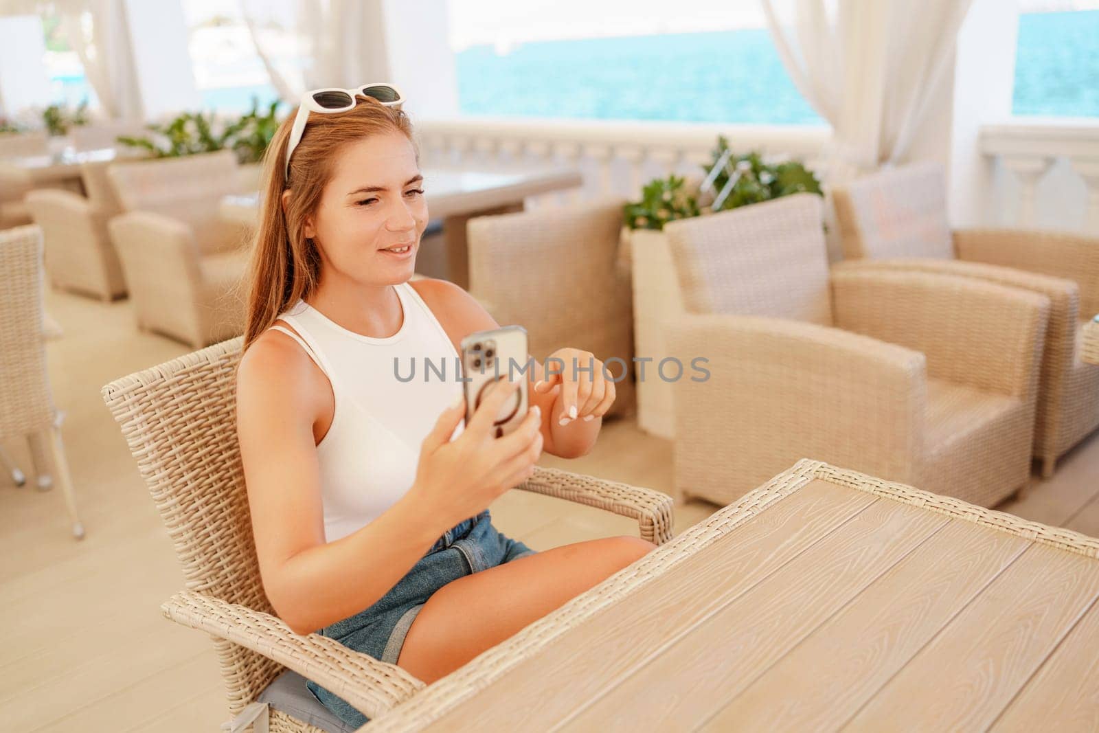 A woman is sitting at a table with a cell phone in her hand. She is taking a picture of the ocean. by Matiunina