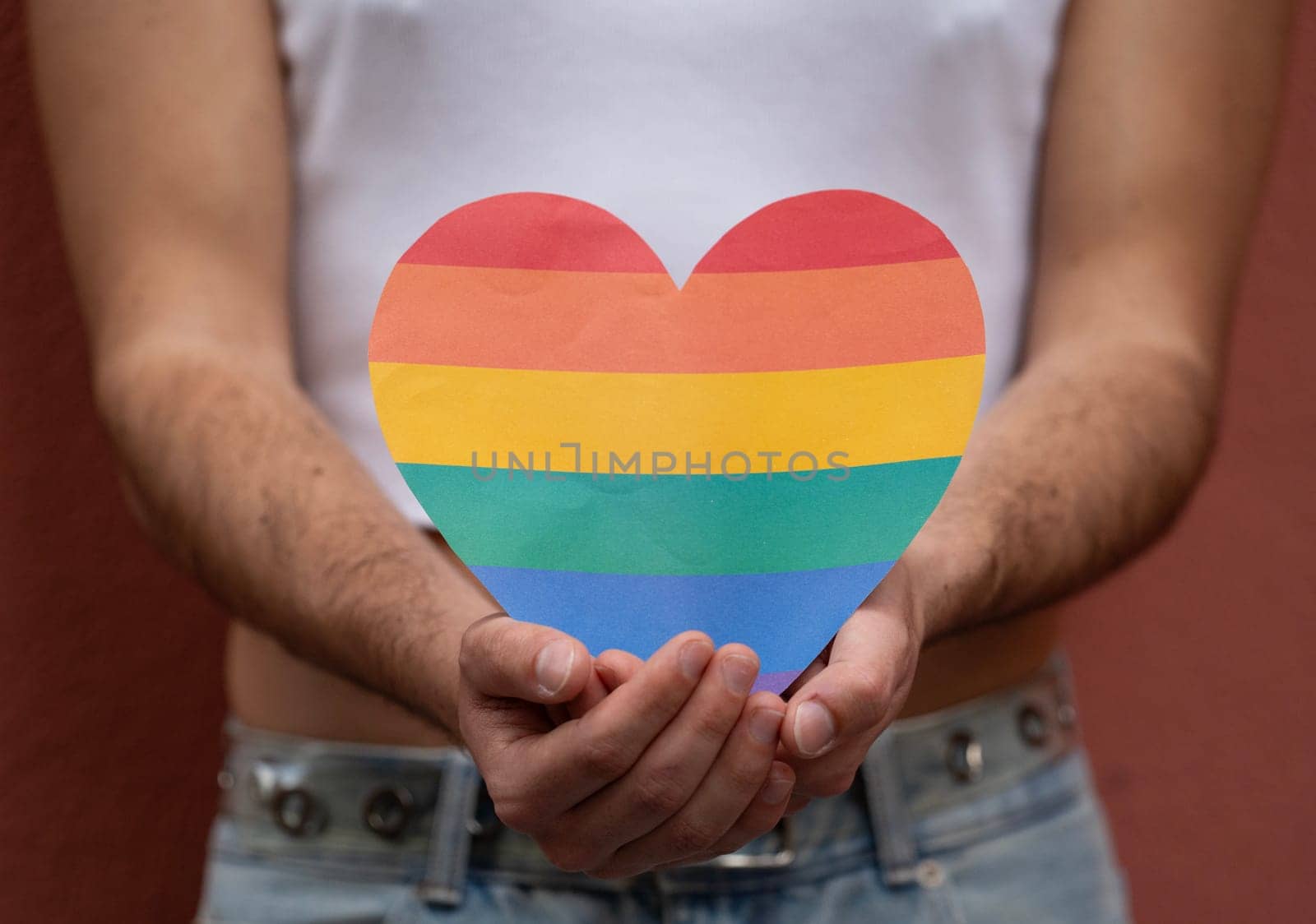 An unrecognizable gay male is holding a rainbow heat symbol with both hands beside his body to support LGBTQ community.