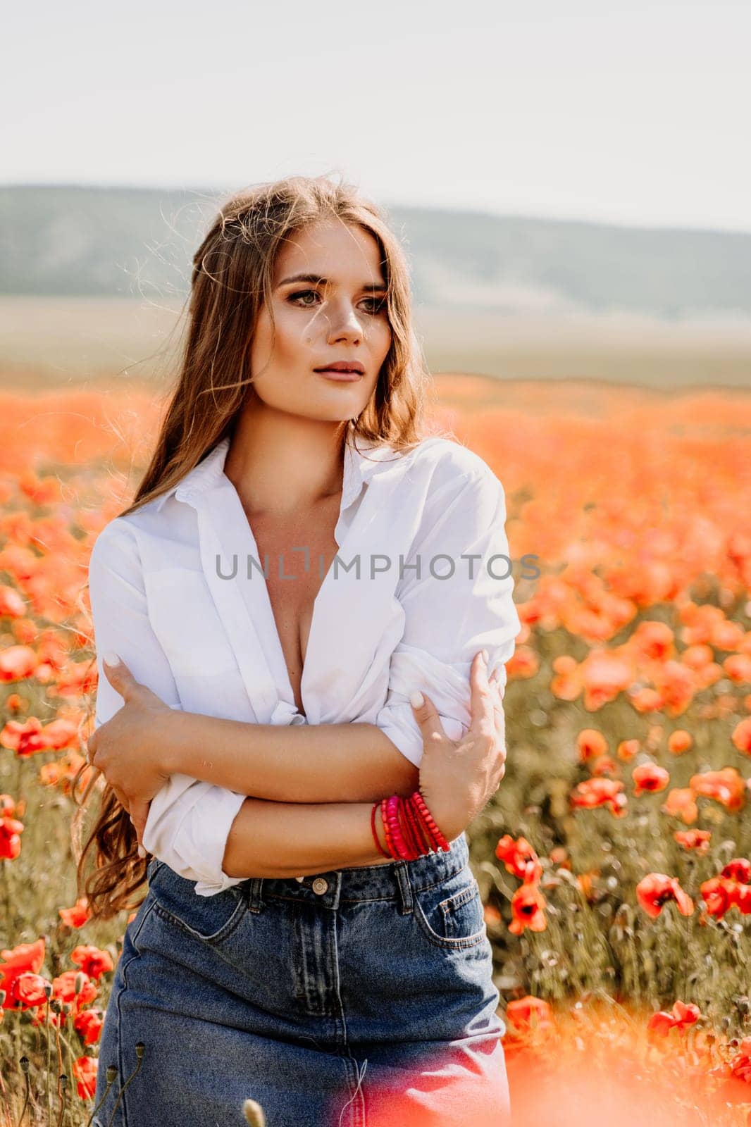 Happy woman in a poppy field in a white shirt and denim skirt with a wreath of poppies on her head posing and enjoying the poppy field