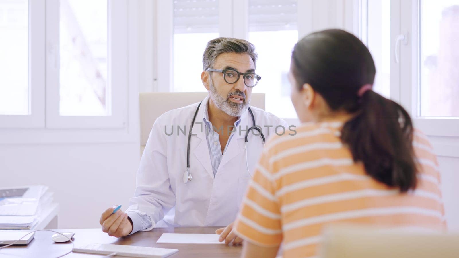 Mature doctor writing down notes and talking to a female patient sitting in a clinic