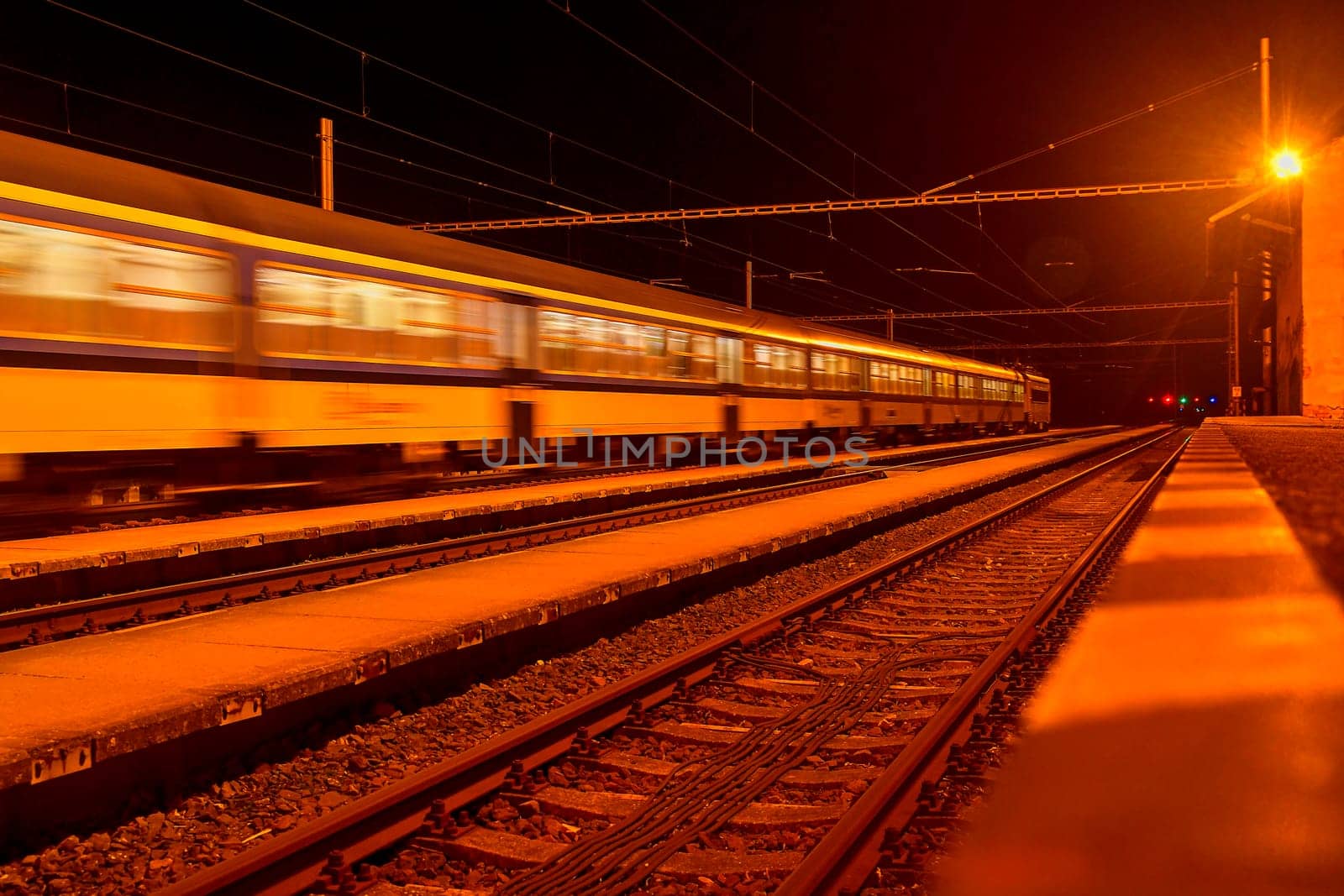 White and blue train passing a small roofless train station at night in the Czech Republic. Blurred motion train.