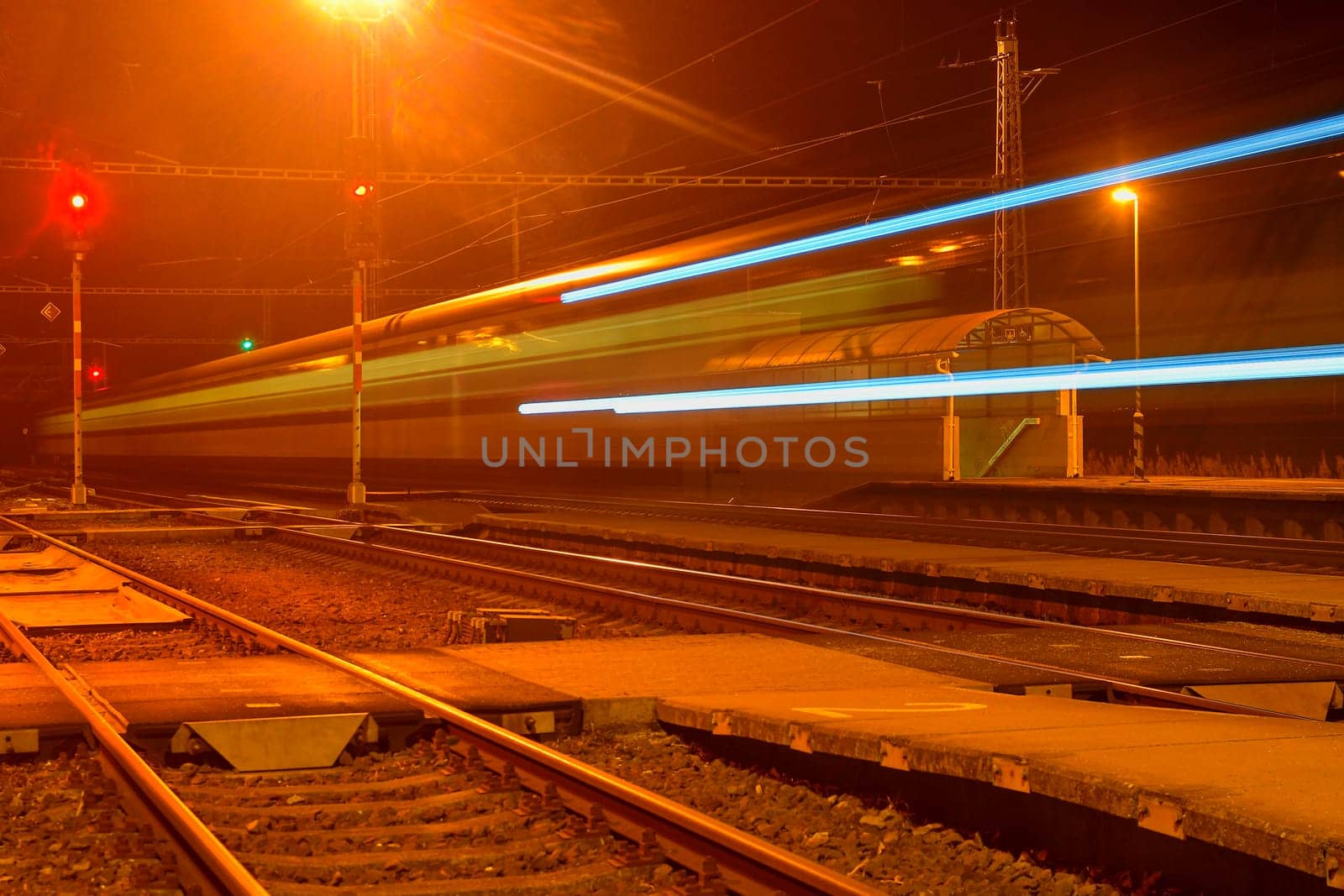White and blue train passing a small roofless train station at night in the Czech Republic. Blurred motion train by roman_nerud