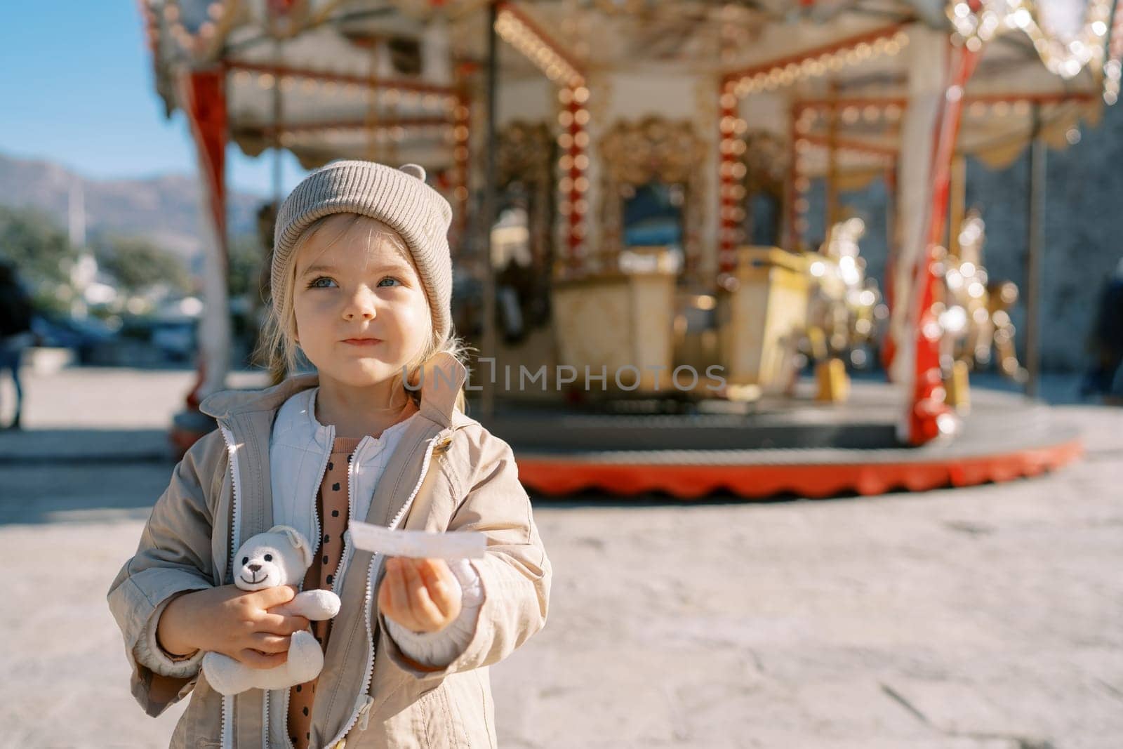 Little girl with a teddy bear stands with a ticket in her hand near a spinning carousel. High quality photo