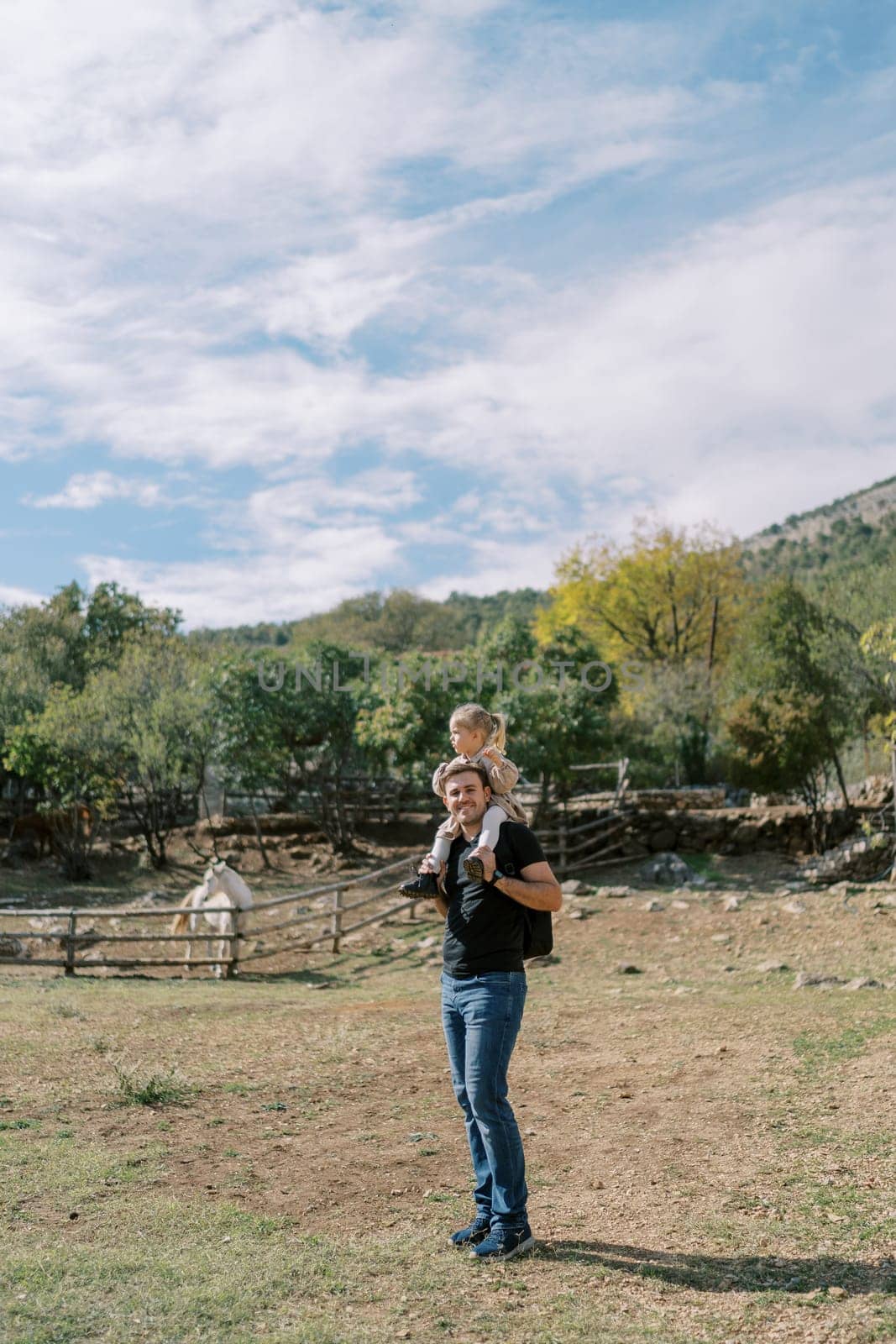 Dad with a little girl on his shoulders stands near a paddock with a horse in the park. High quality photo