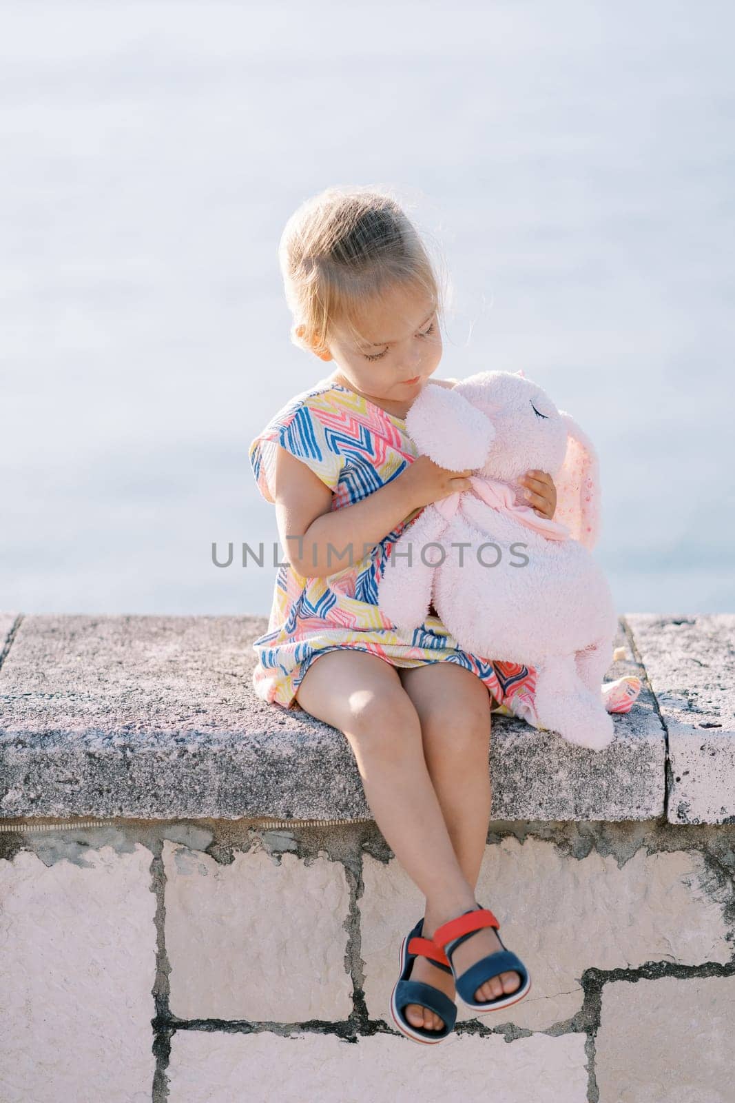 Little girl examines a plush rabbit in her hands while sitting on a fence by the sea. High quality photo