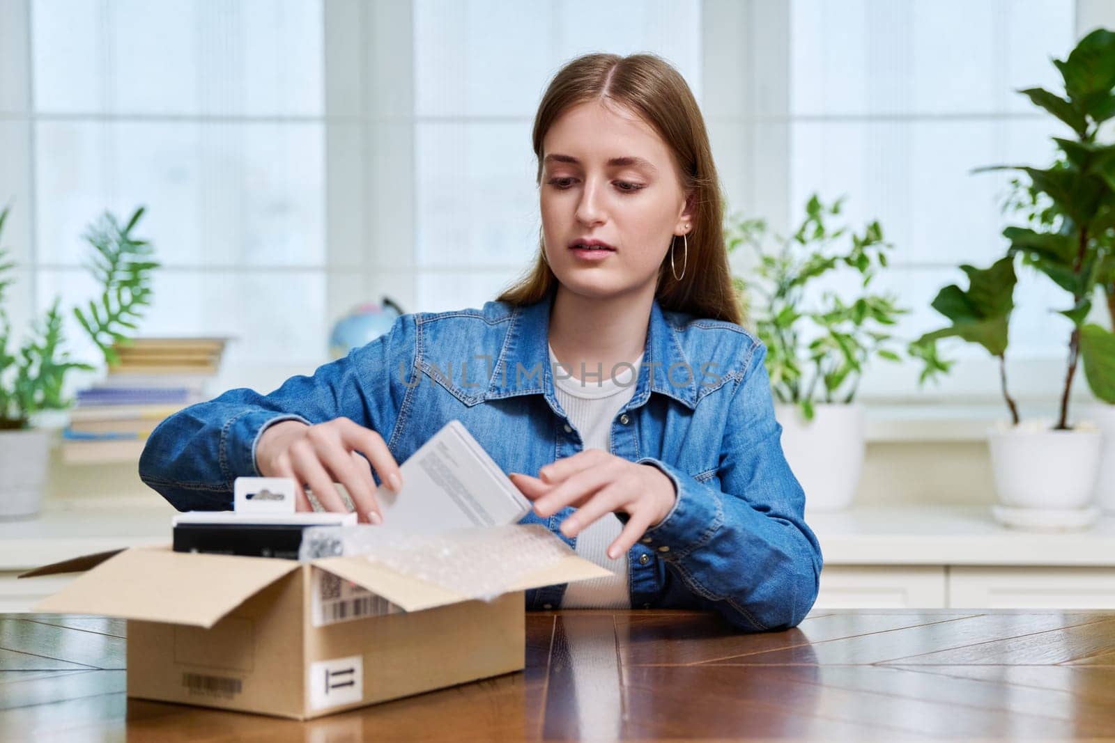 Satisfied young teen female shopper consumer sits at home and unpacks cardboard box with online purchases. Teenager girl unpacking box with new smartphone, headphones. Delivery by mail, online store