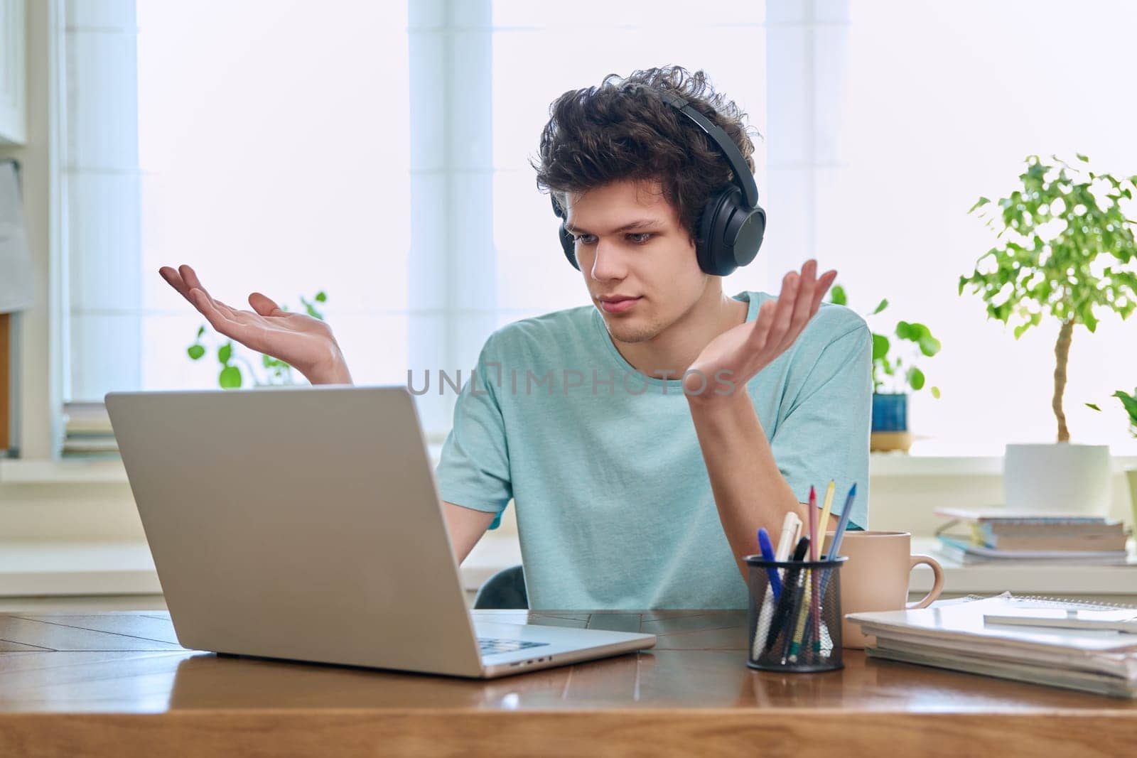 Young male college student sitting at desk, talking in web camera of laptop by VH-studio