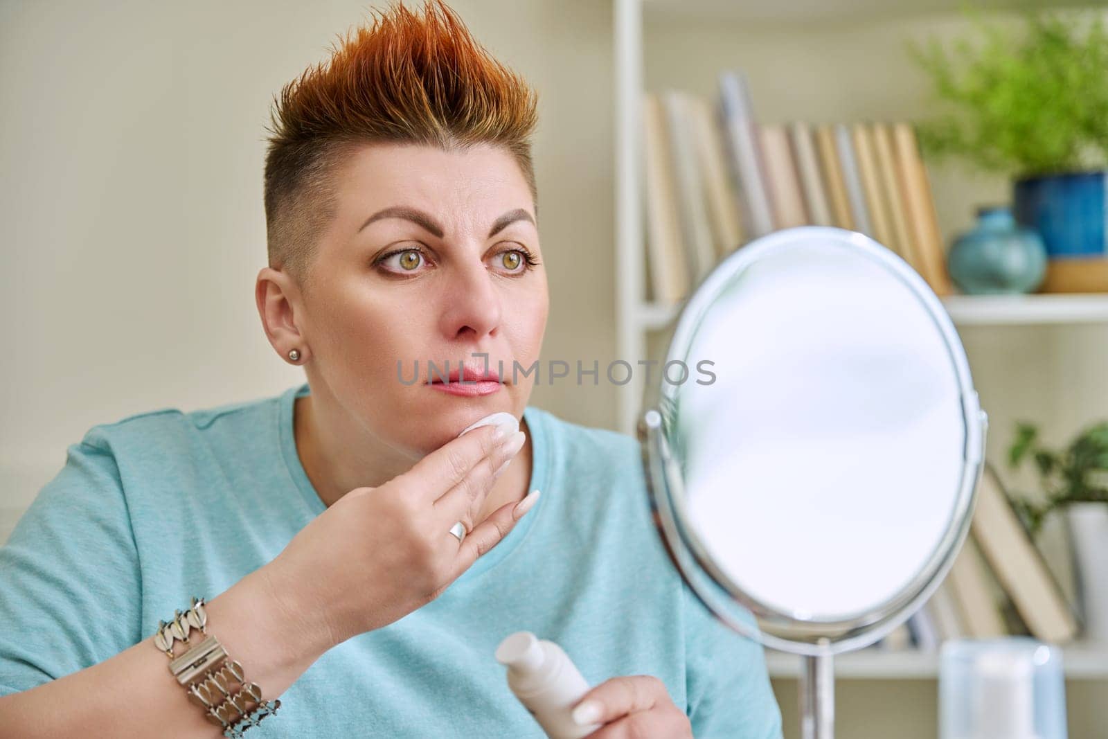 Middle-aged woman with a mirror taking care of her facial skin by VH-studio
