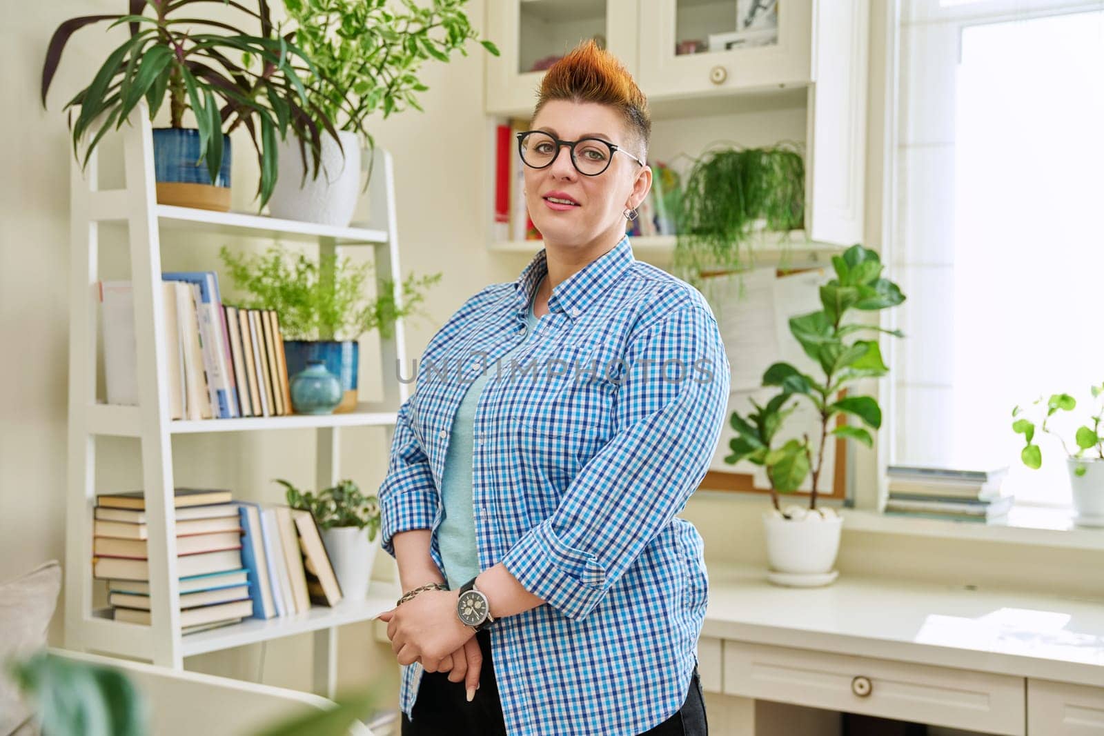 Portrait of smiling middle-aged woman in glasses with red haircut looking at camera in home interior. Mature people, lifestyle, health, life concept