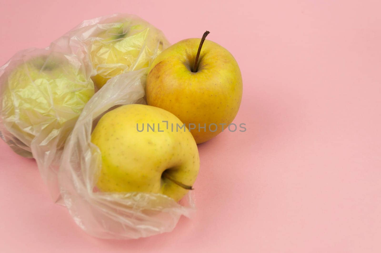 A bag of apples is sitting on a pink background. The apples are in plastic bags and are yellow in color