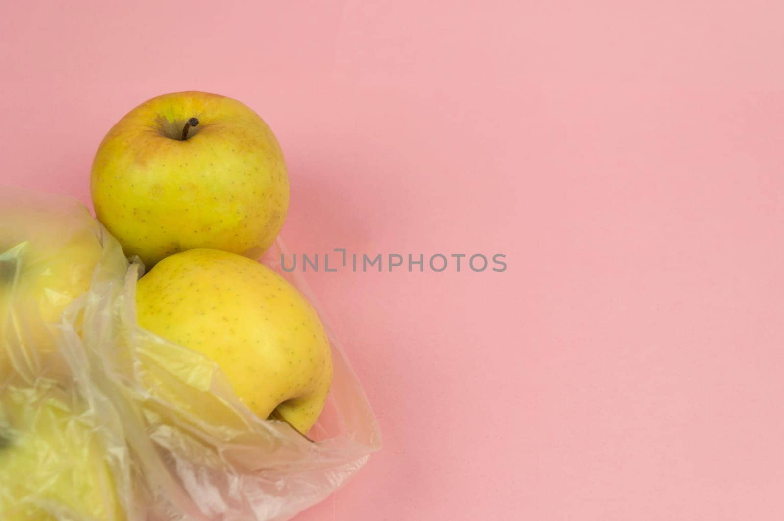 Three apples in a plastic bag on a pink background. The apples are yellow and shiny